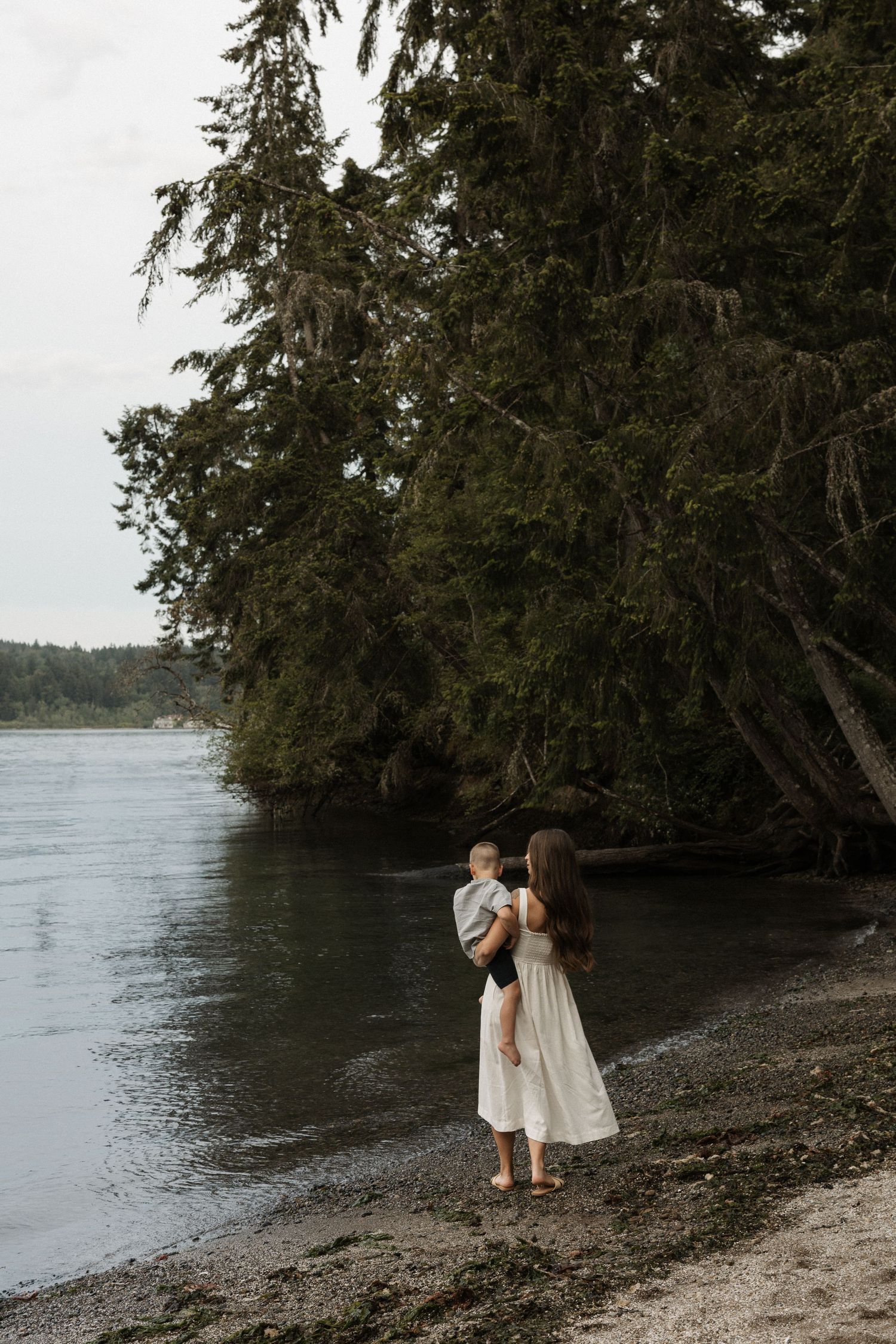 A mother in a white dress holds her baby on a peaceful rocky shoreline surrounded by tall evergreen trees.