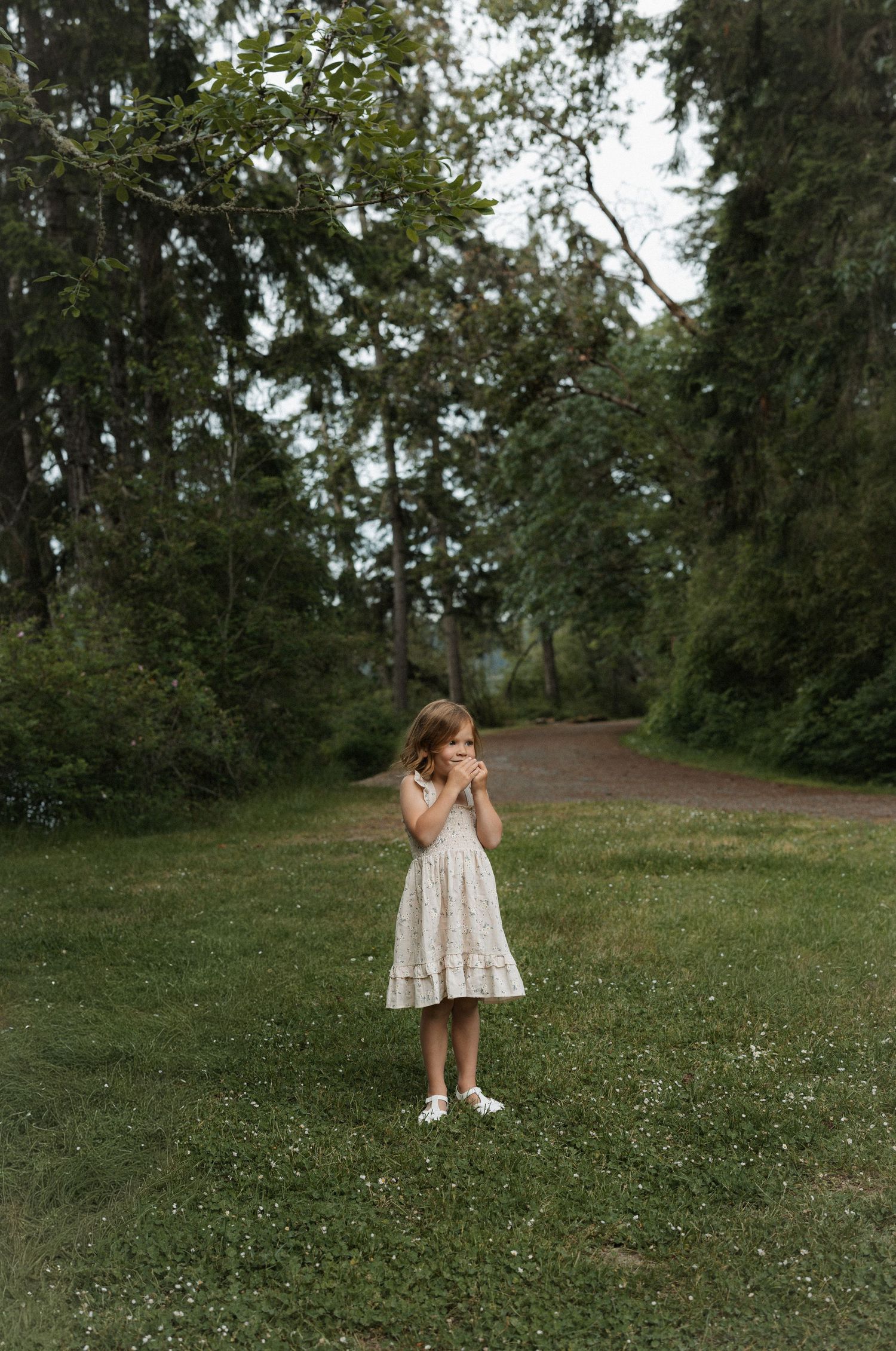 A young girl in a floral dress stands on green grass surrounded by lush trees in a serene outdoor setting.