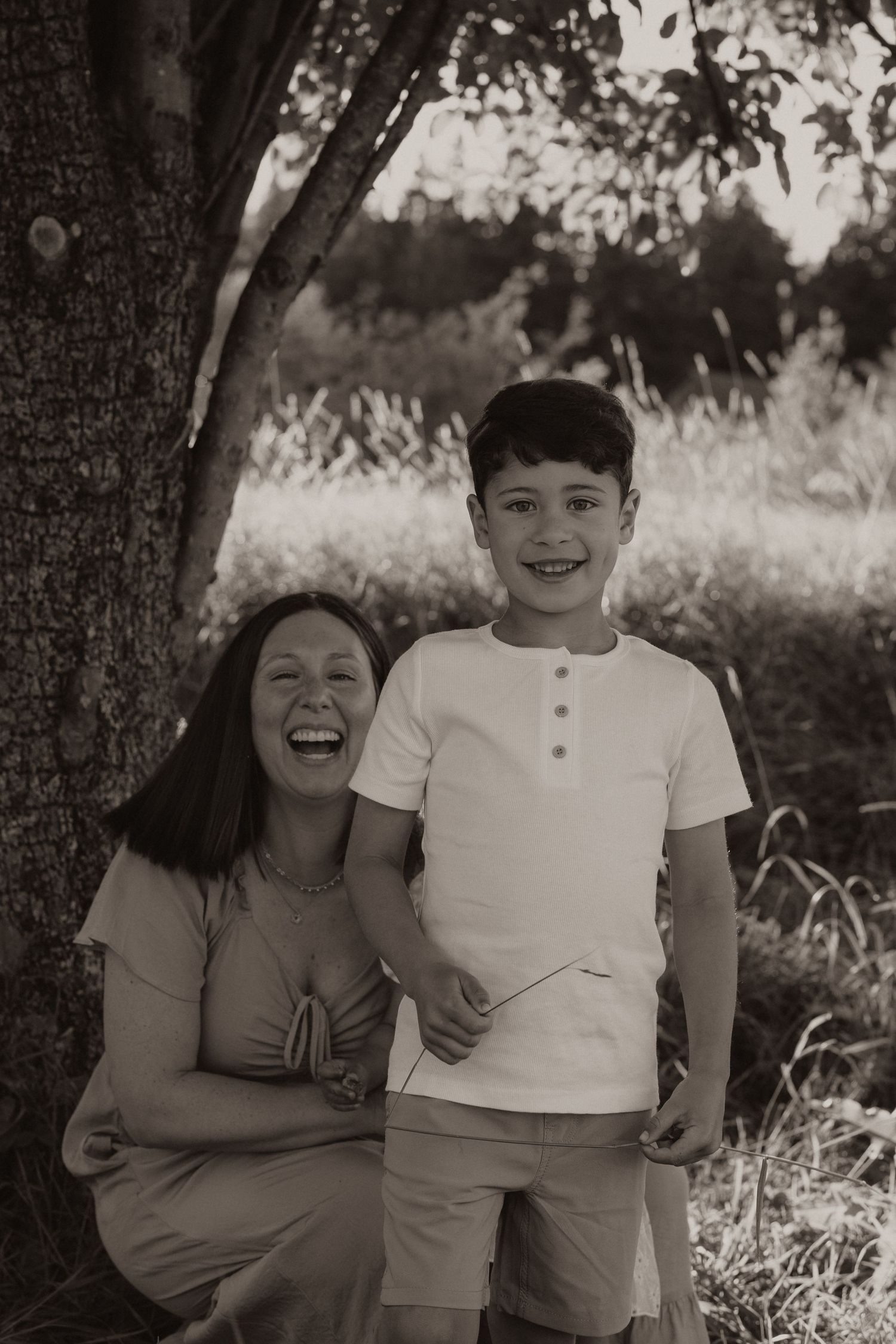 Black and white photo of laughing mother and smiling son beside a tree outdoors.