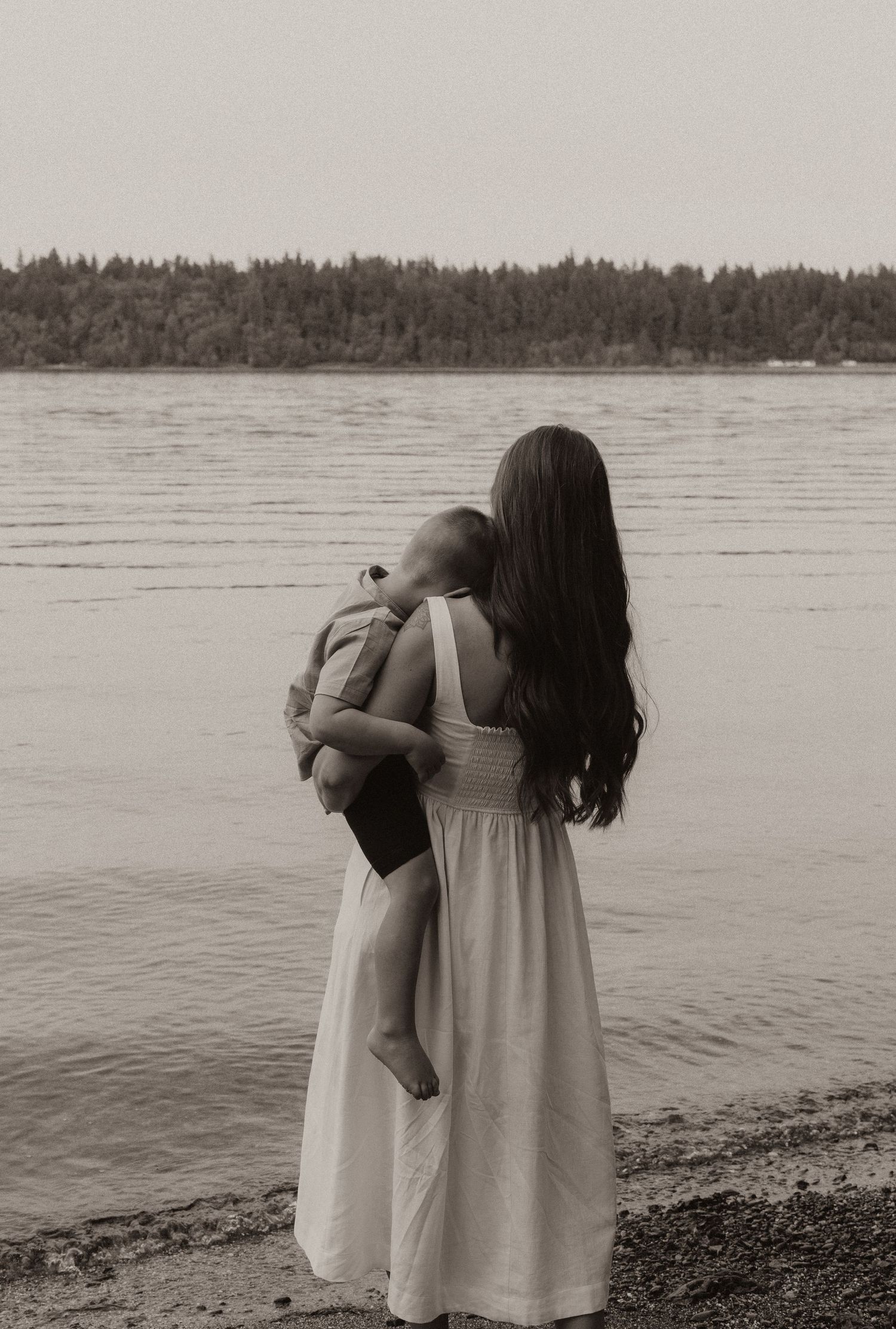 Mother in white dress holding baby by a serene lake with forested shoreline in black and white.