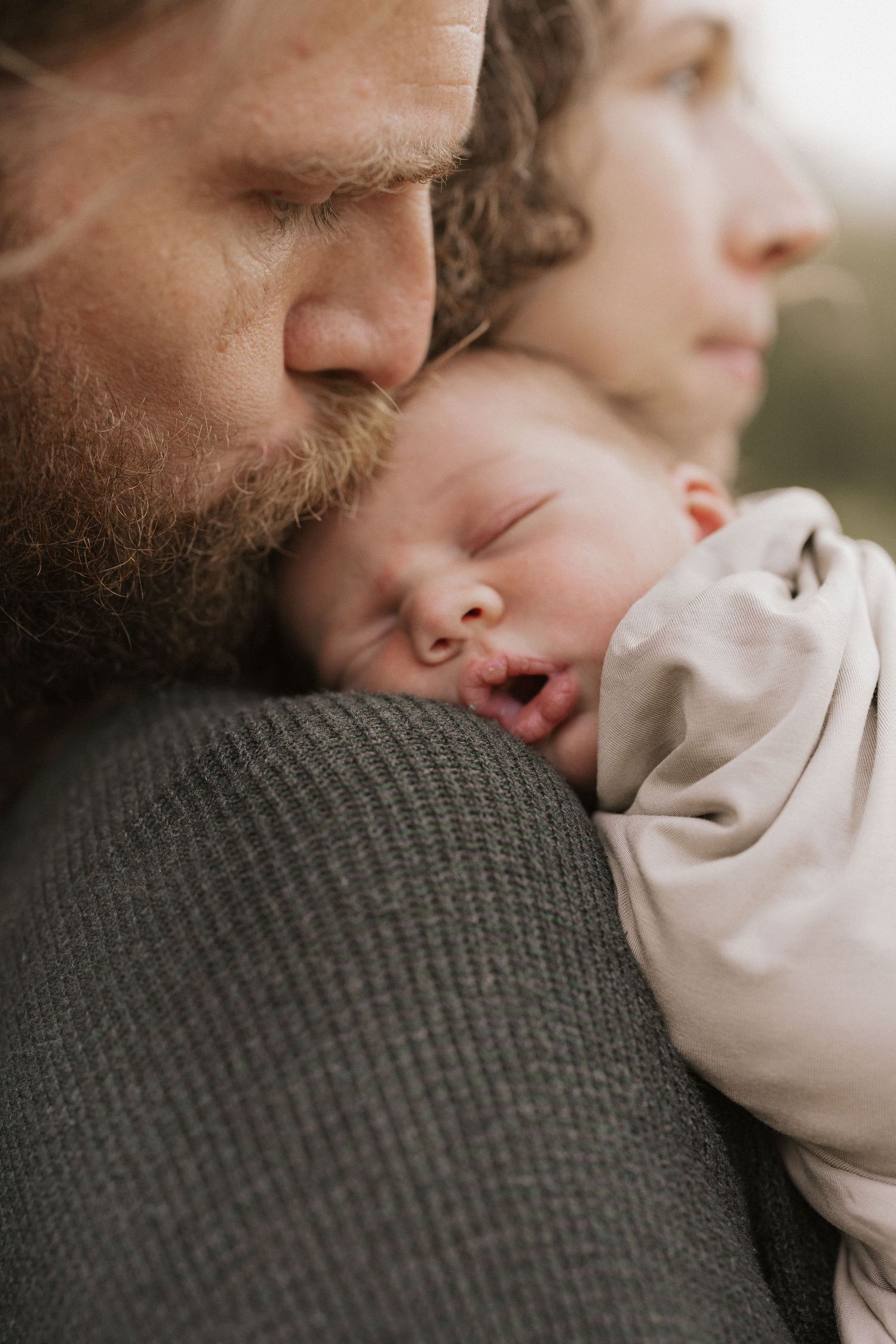 A bearded father tenderly kisses his sleeping newborn baby held against his chest.