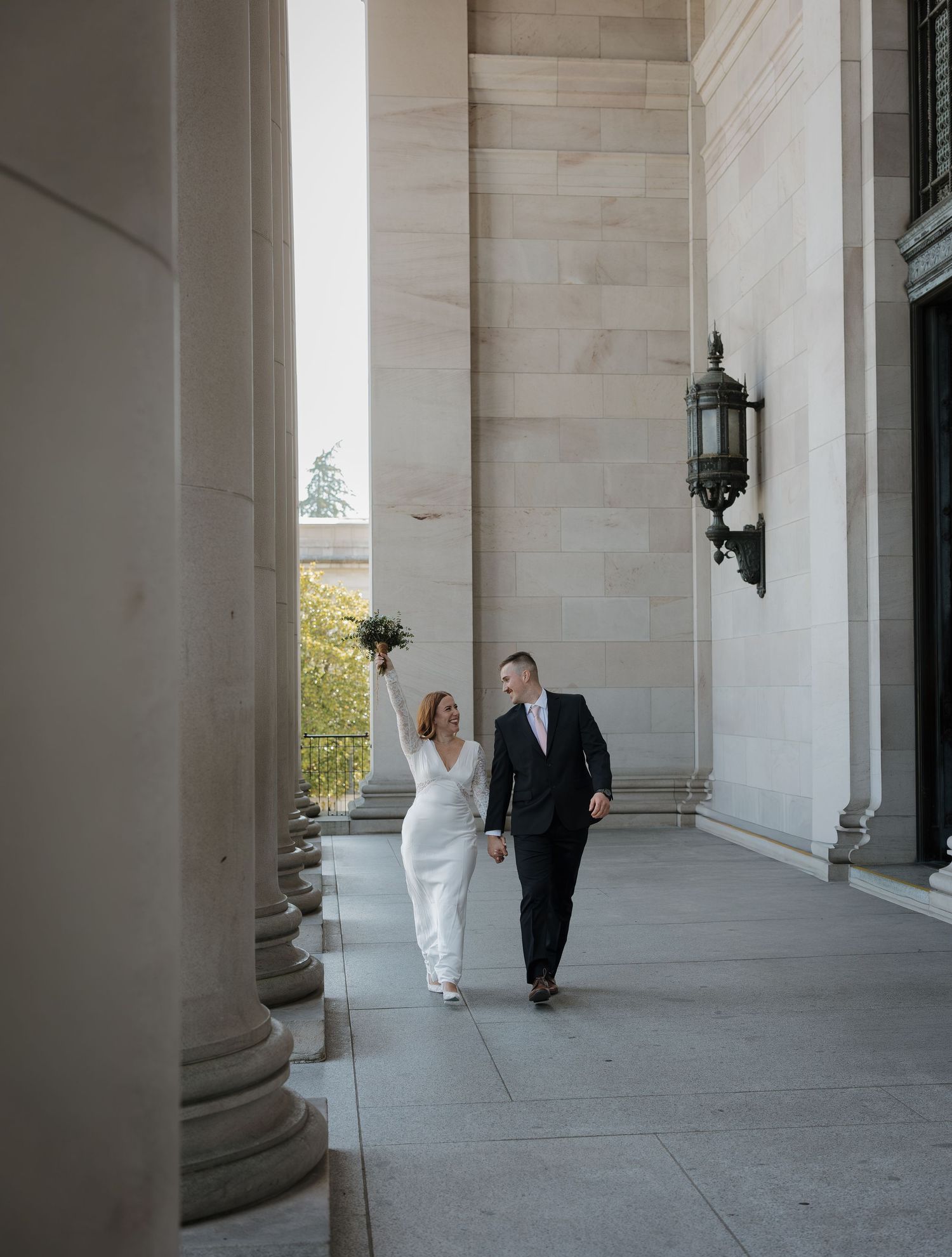 A bride and groom walk hand-in-hand along a grand columned building exterior on their wedding day.
