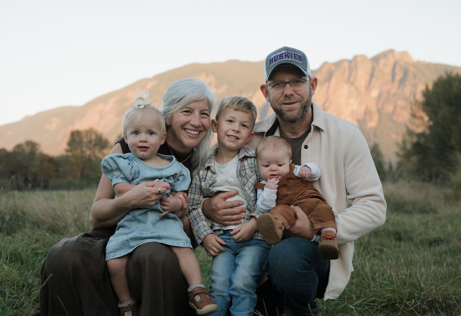 Family of five poses outdoors with mountain backdrop at sunset, parents holding three young children in a green meadow.