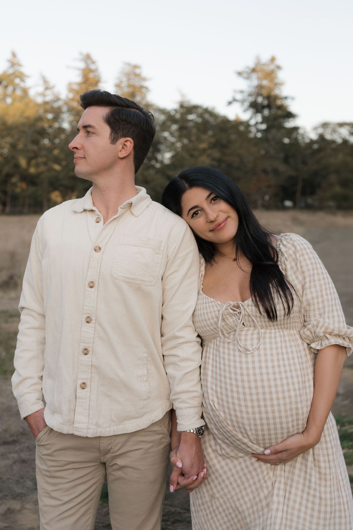 Expecting couple in matching cream outfits stand close together holding hands in a golden field during an outdoor maternity photo session.