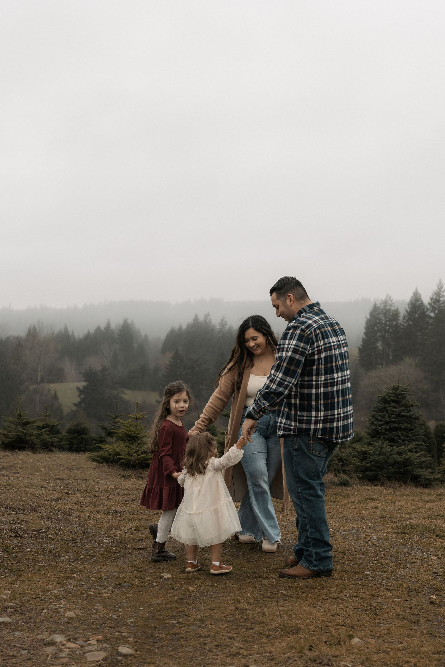 A family of four poses on a misty hilltop surrounded by evergreen trees, with parents holding hands with two young girls.