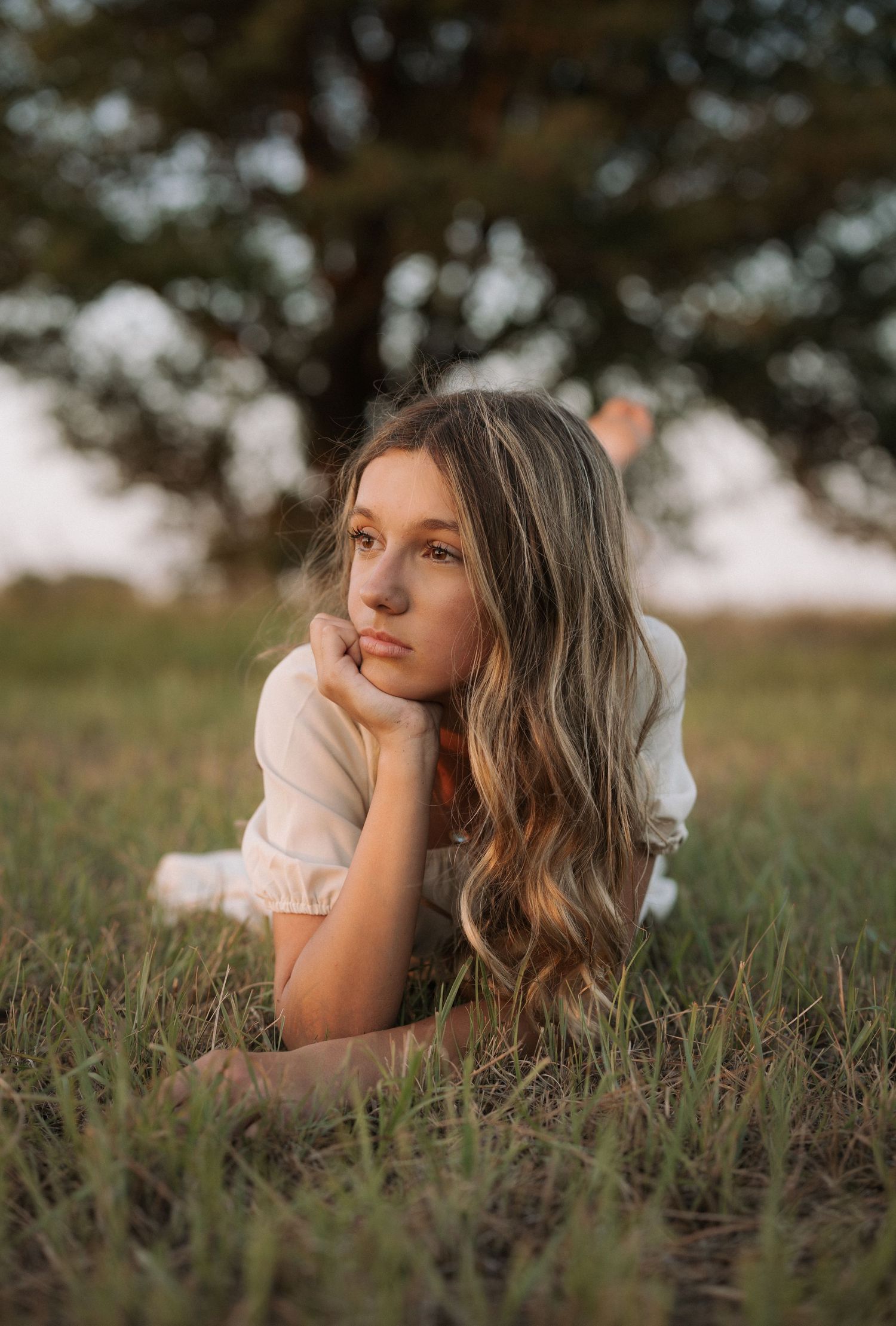 Young woman with long wavy hair lying in grass, resting chin on hands, wearing white top with tree backdrop.