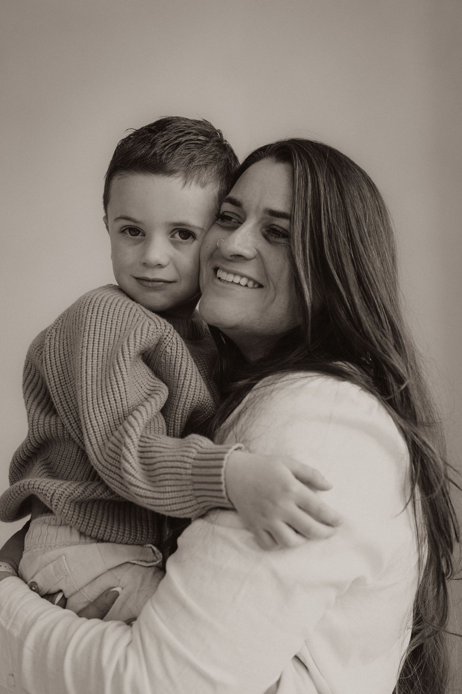 Black and white portrait of smiling mother hugging young boy in knit sweater against neutral background.