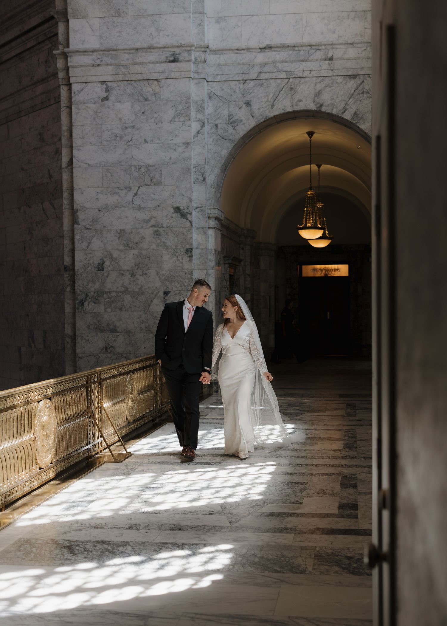 Bride and groom walk hand in hand through a sunlit marble hallway with arched ceilings at an elegant venue.
