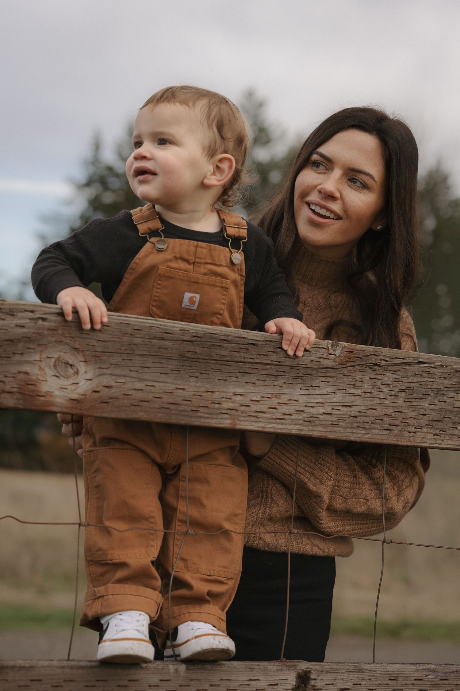 A smiling woman and toddler in brown Carhartt overalls lean against a wooden fence outdoors.