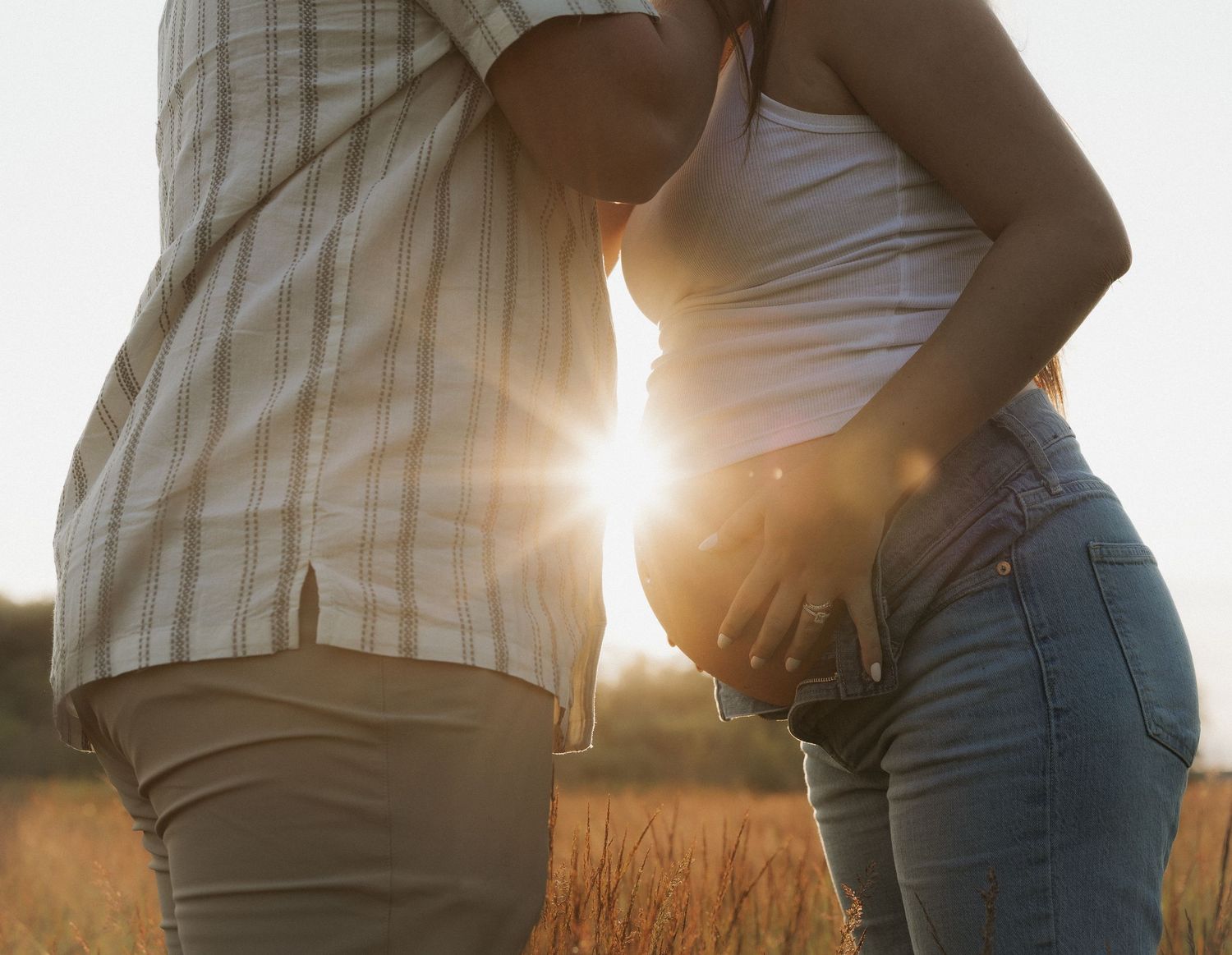 Pregnant couple standing in sunlit field at golden hour, woman cradling baby bump in jeans and white top.