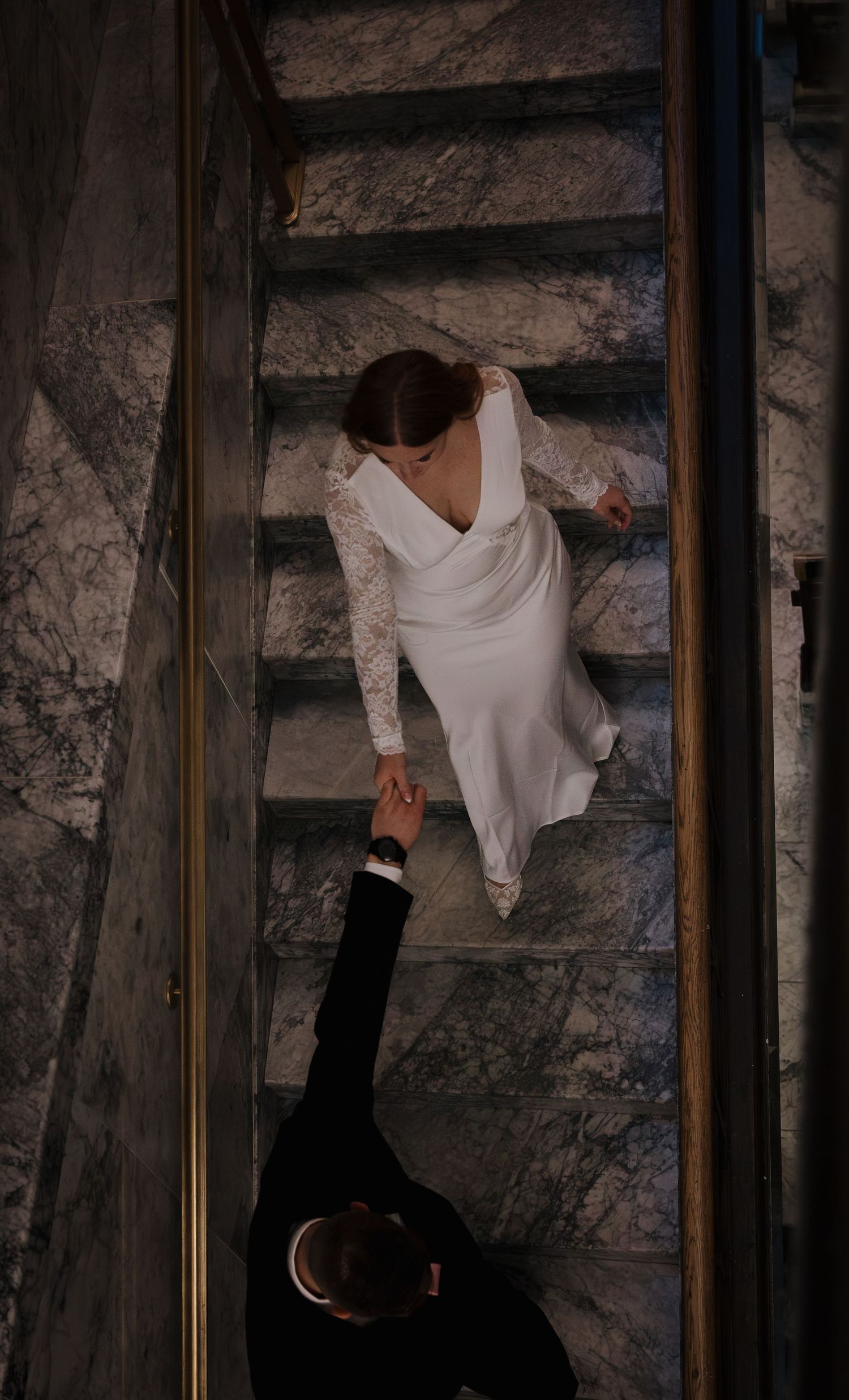 Aerial view of bride in white lace gown holding groom's hand on marble staircase.