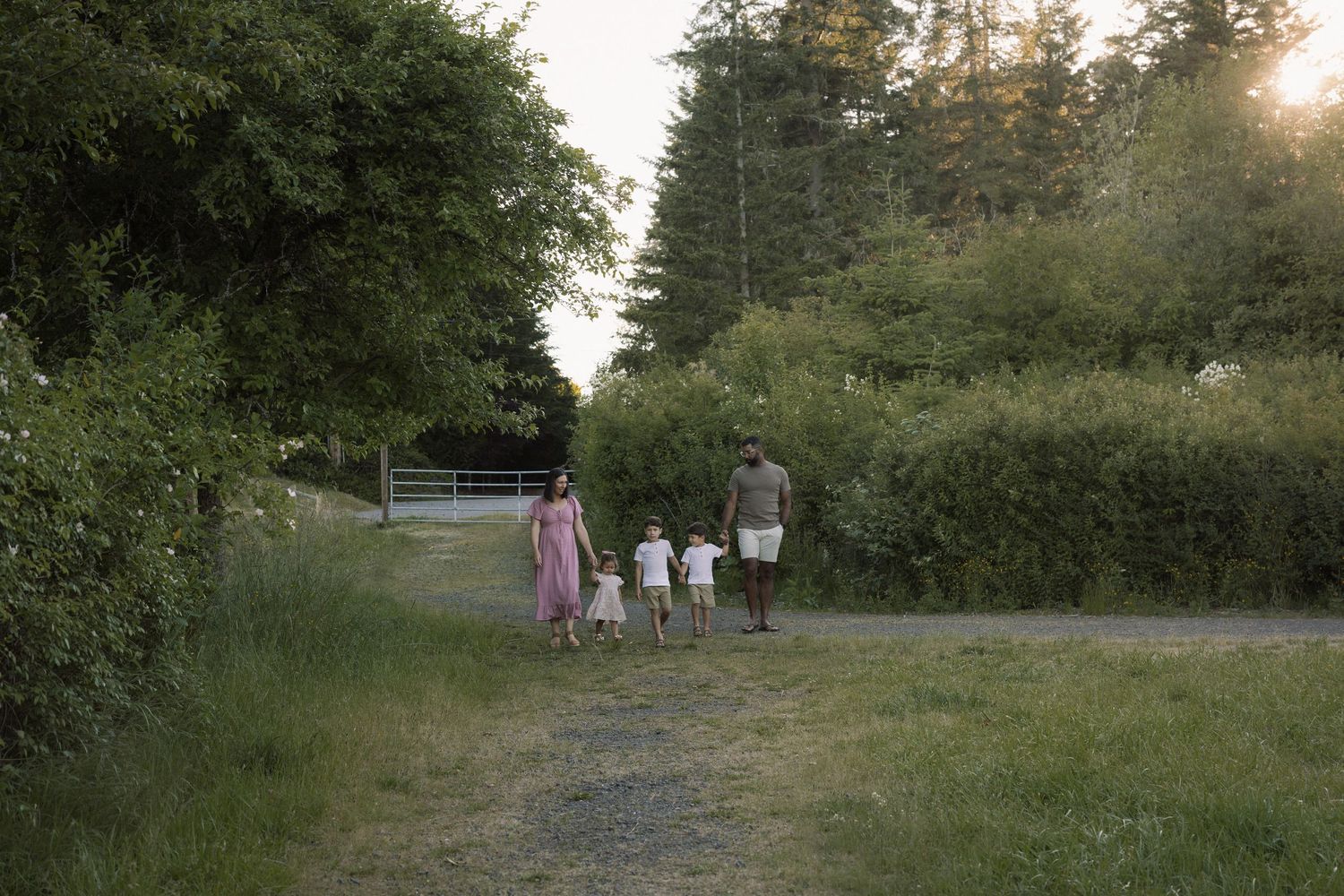 Family walking hand-in-hand down a scenic rural path surrounded by lush green trees at sunset.
