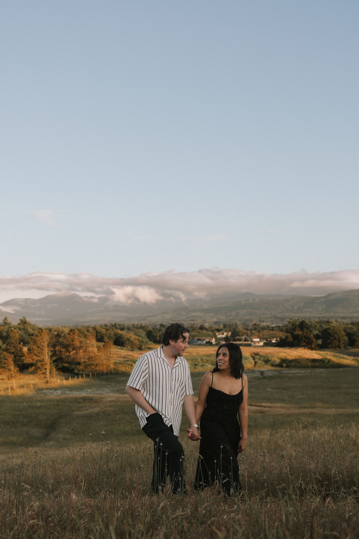 A couple holds hands in a sunlit field with misty mountains in the background.