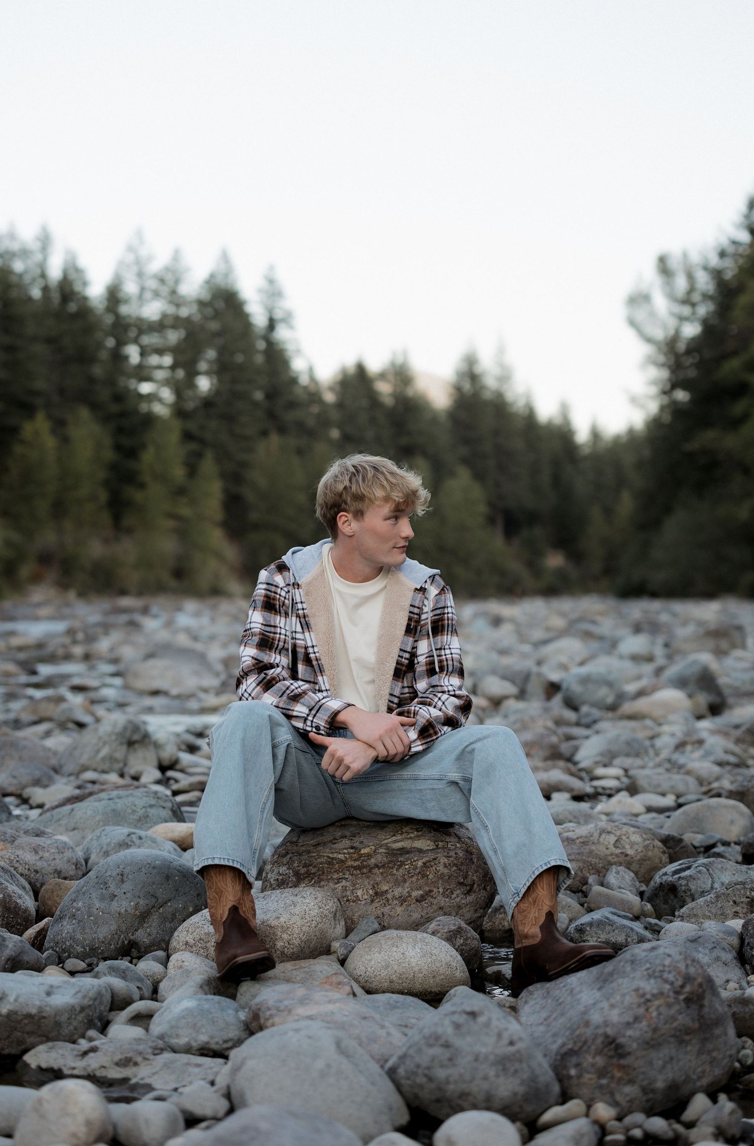 Young blonde man sitting on rocks in a rocky riverbed wearing plaid shirt and jeans with evergreen trees behind him.