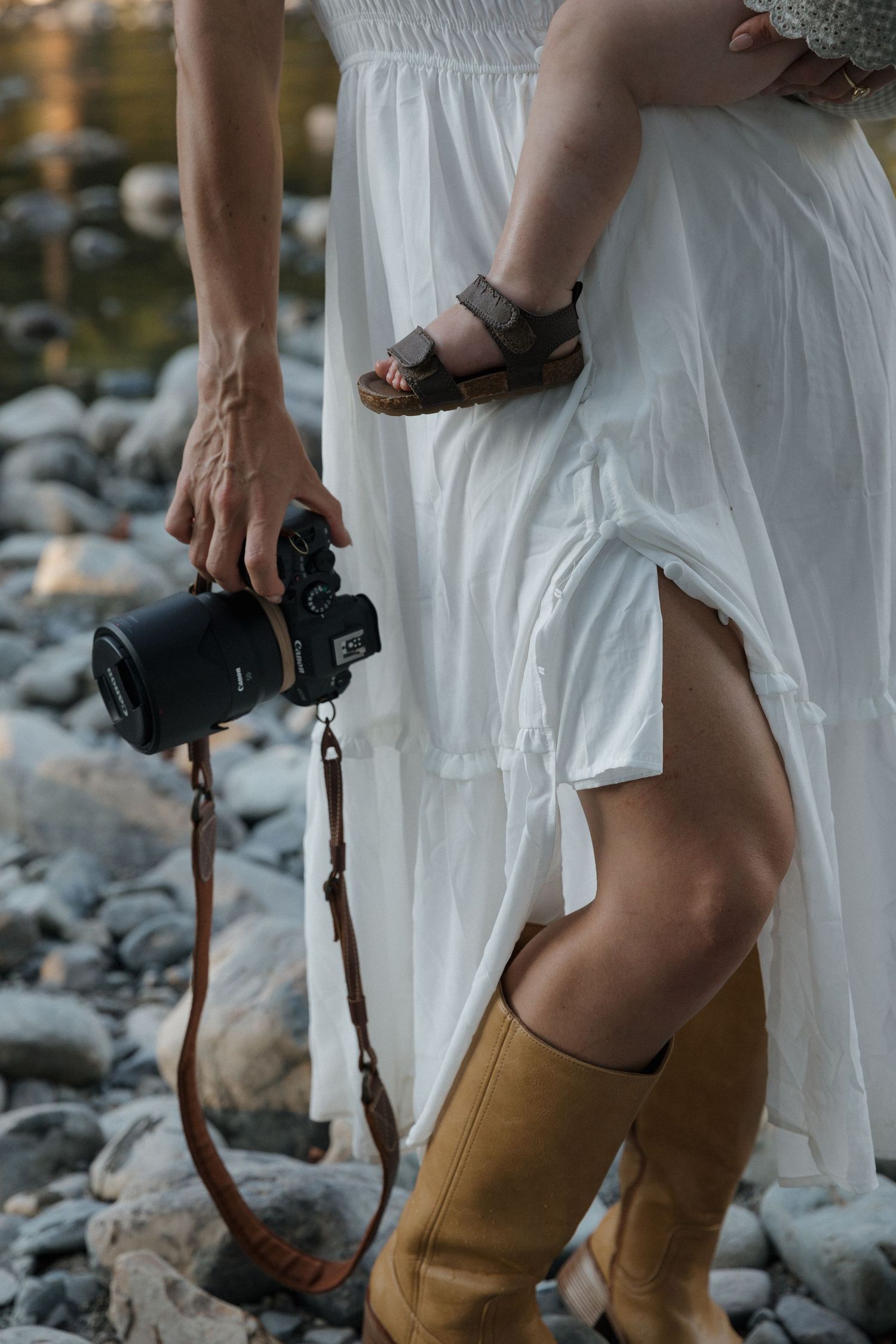 Woman in white dress holding camera and baby on rocky shore, wearing tan knee-high boots outdoors.