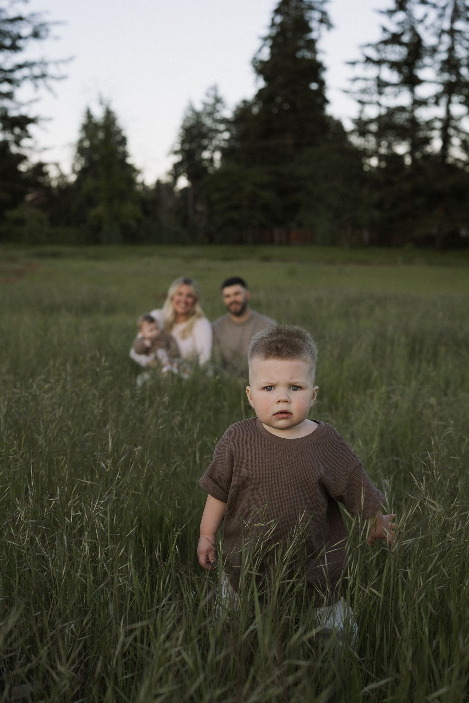 Toddler in brown shirt stands in tall green grass as parents smile in background during family photo session.