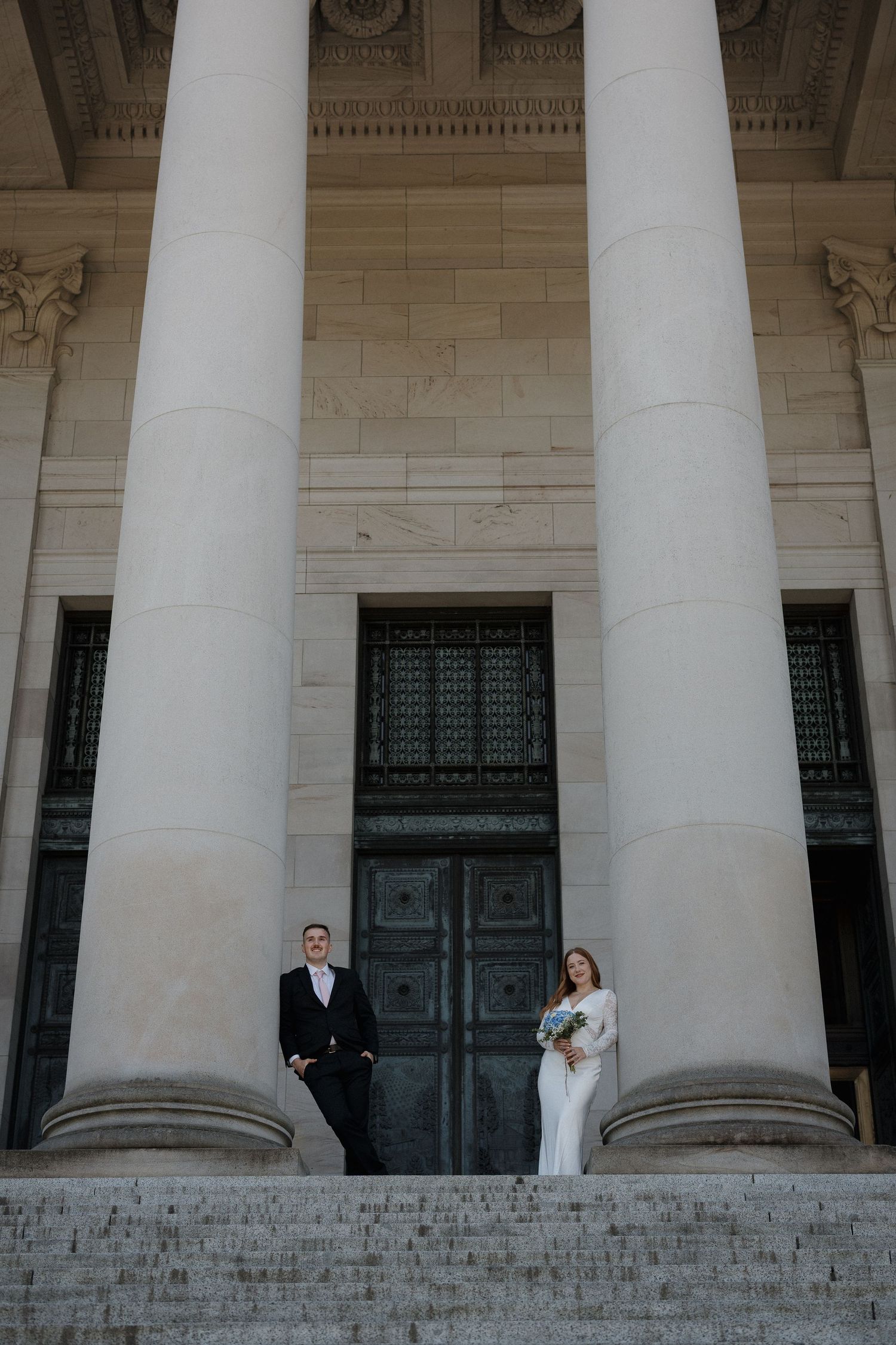 Bride and groom posing between grand columns on steps of a neoclassical building on their wedding day.
