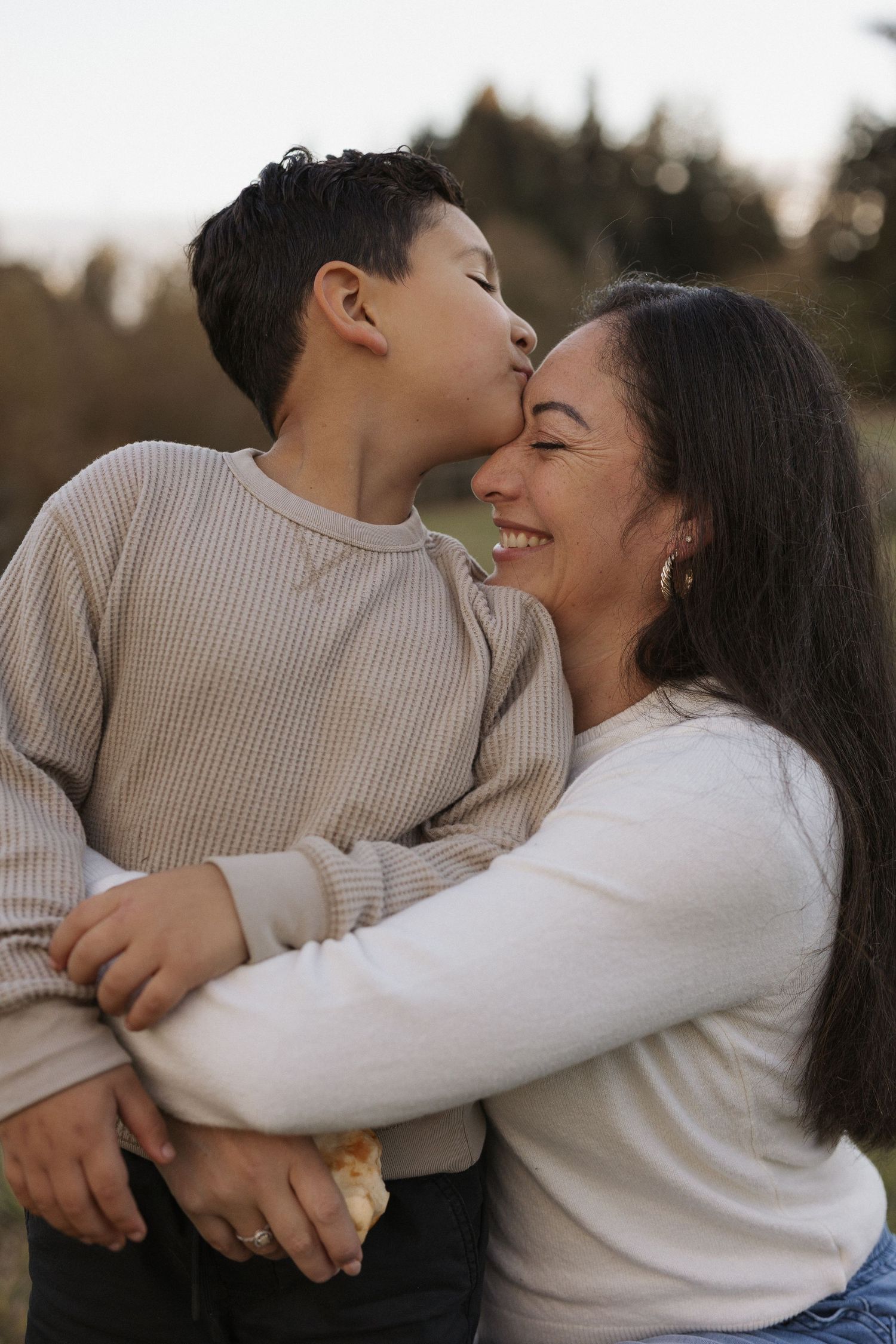 A young boy kisses his smiling mother on the cheek while embracing her outdoors in a warm, candid family moment.