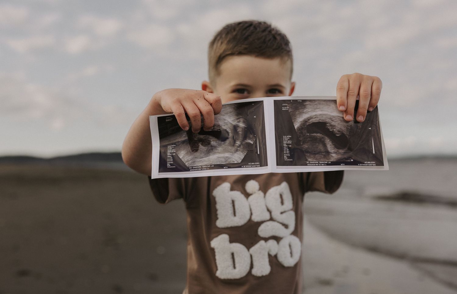 Young boy in big bro shirt holding up ultrasound photos on a beach for a pregnancy announcement.