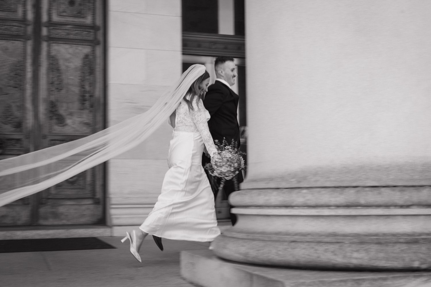 Black and white photo of a bride in a flowing veil and groom walking up steps at a classic wedding venue.