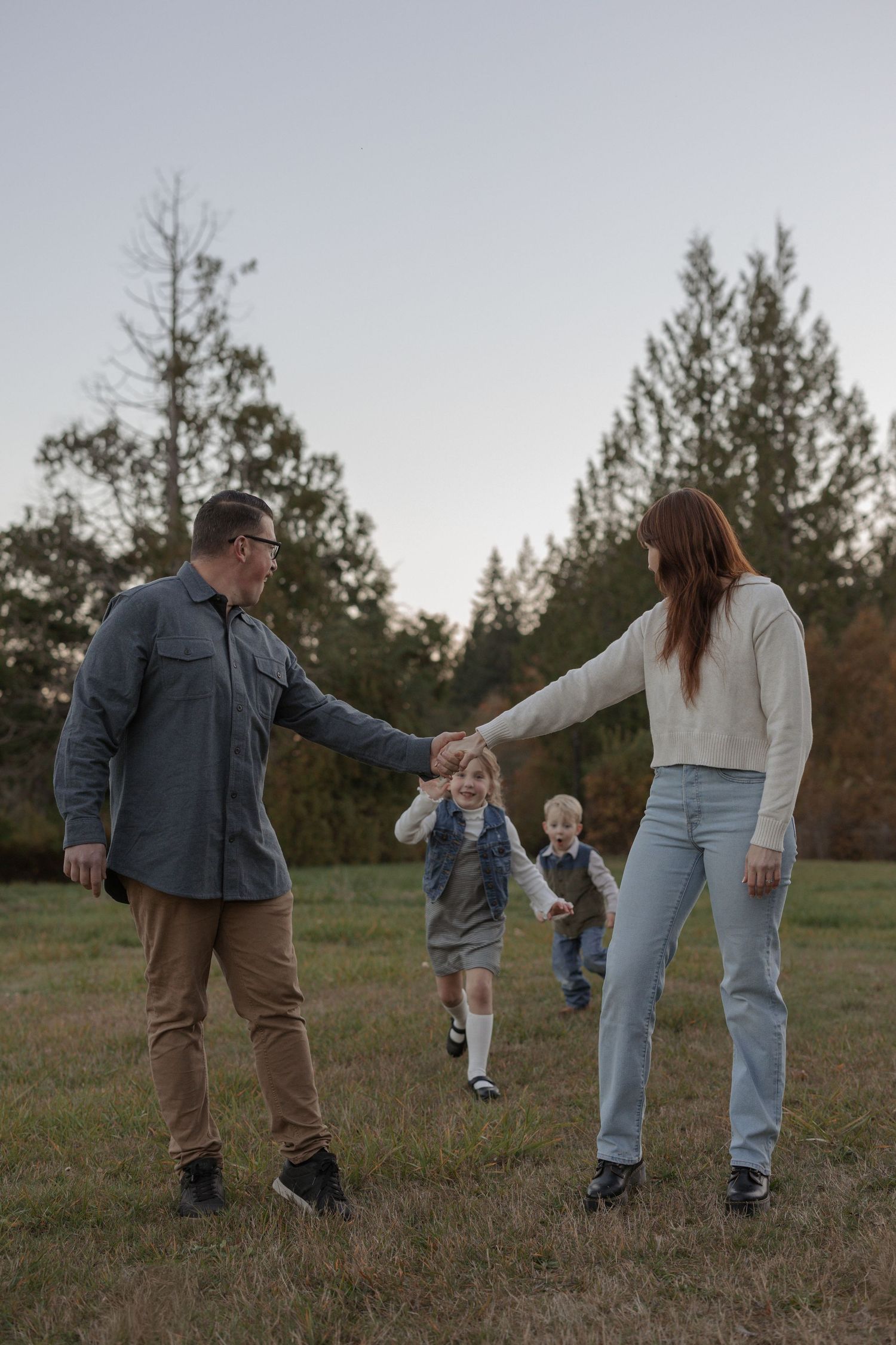 Family of four holding hands while walking in a grassy field surrounded by trees at dusk.