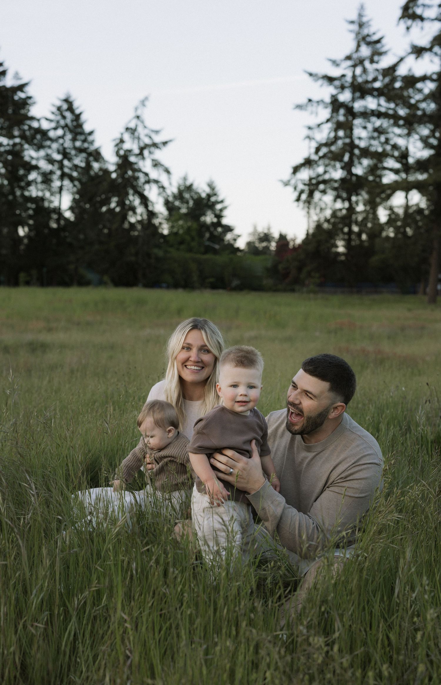 A happy young family sitting in a lush green field with tall trees, parents smiling with their baby toddler outdoors.