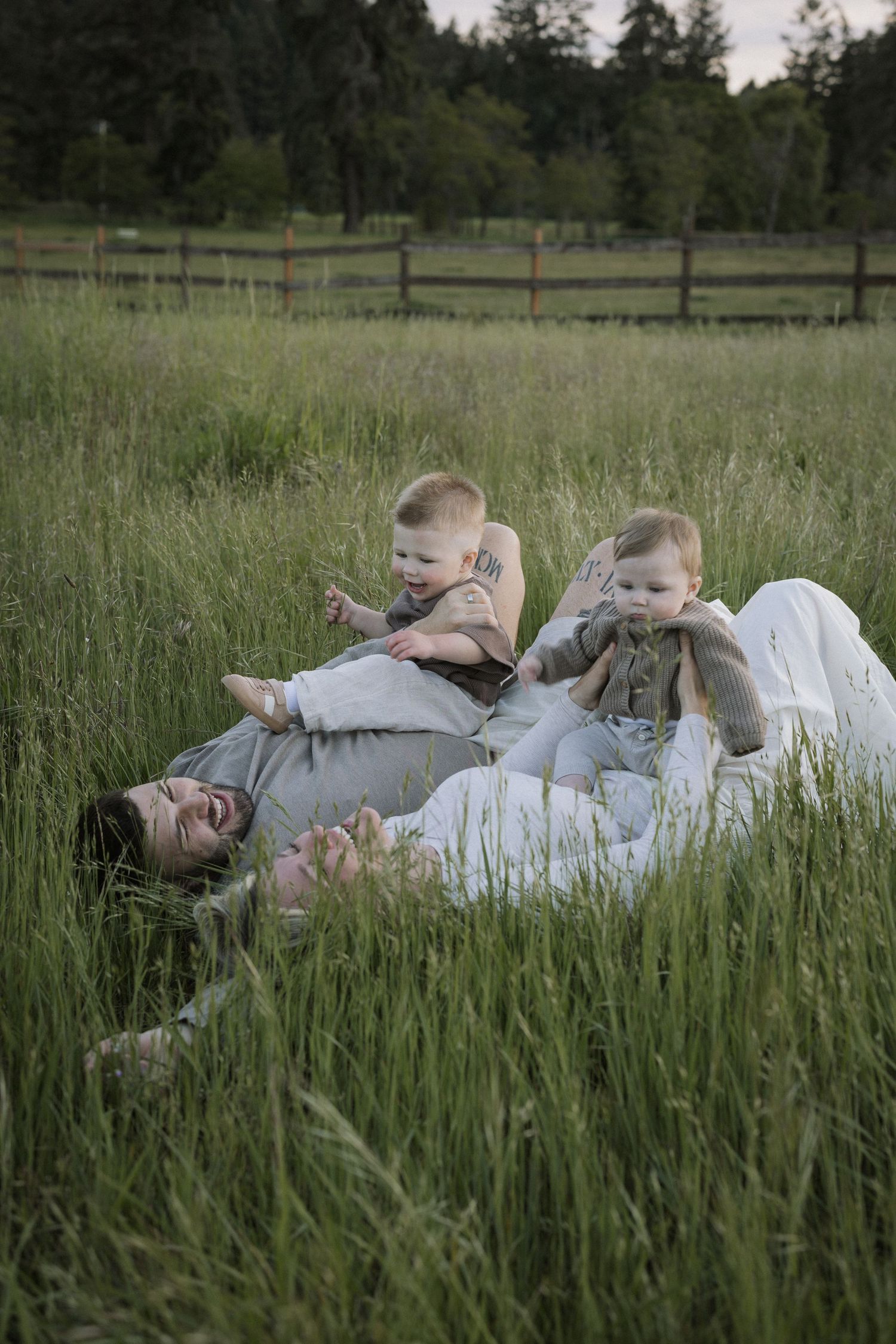 Parent lying in tall green grass with two happy babies sitting on their chest, enjoying a playful outdoor family moment.