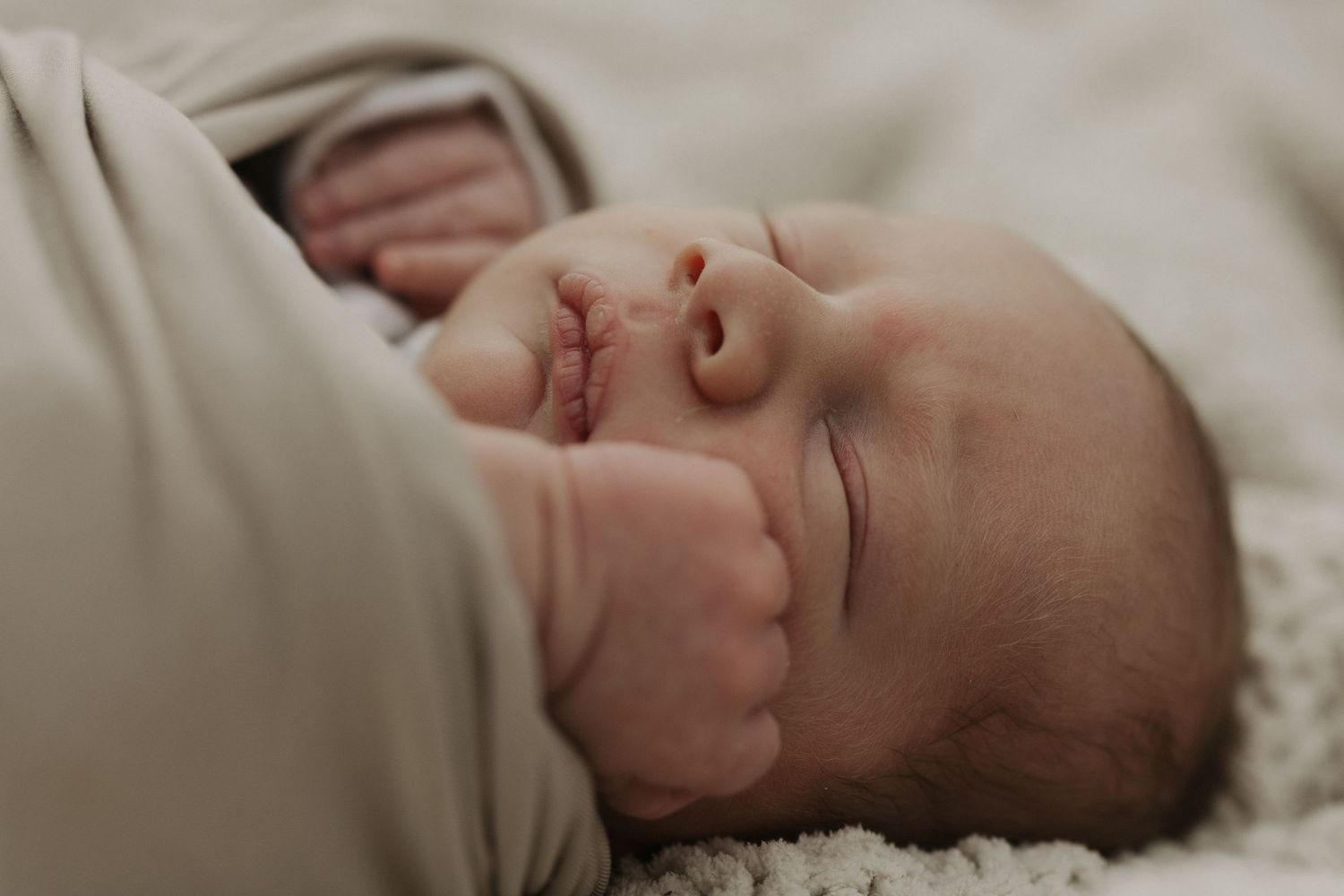 Peaceful newborn baby sleeping swaddled in soft cream blanket, eyes closed, tiny fist raised near face.