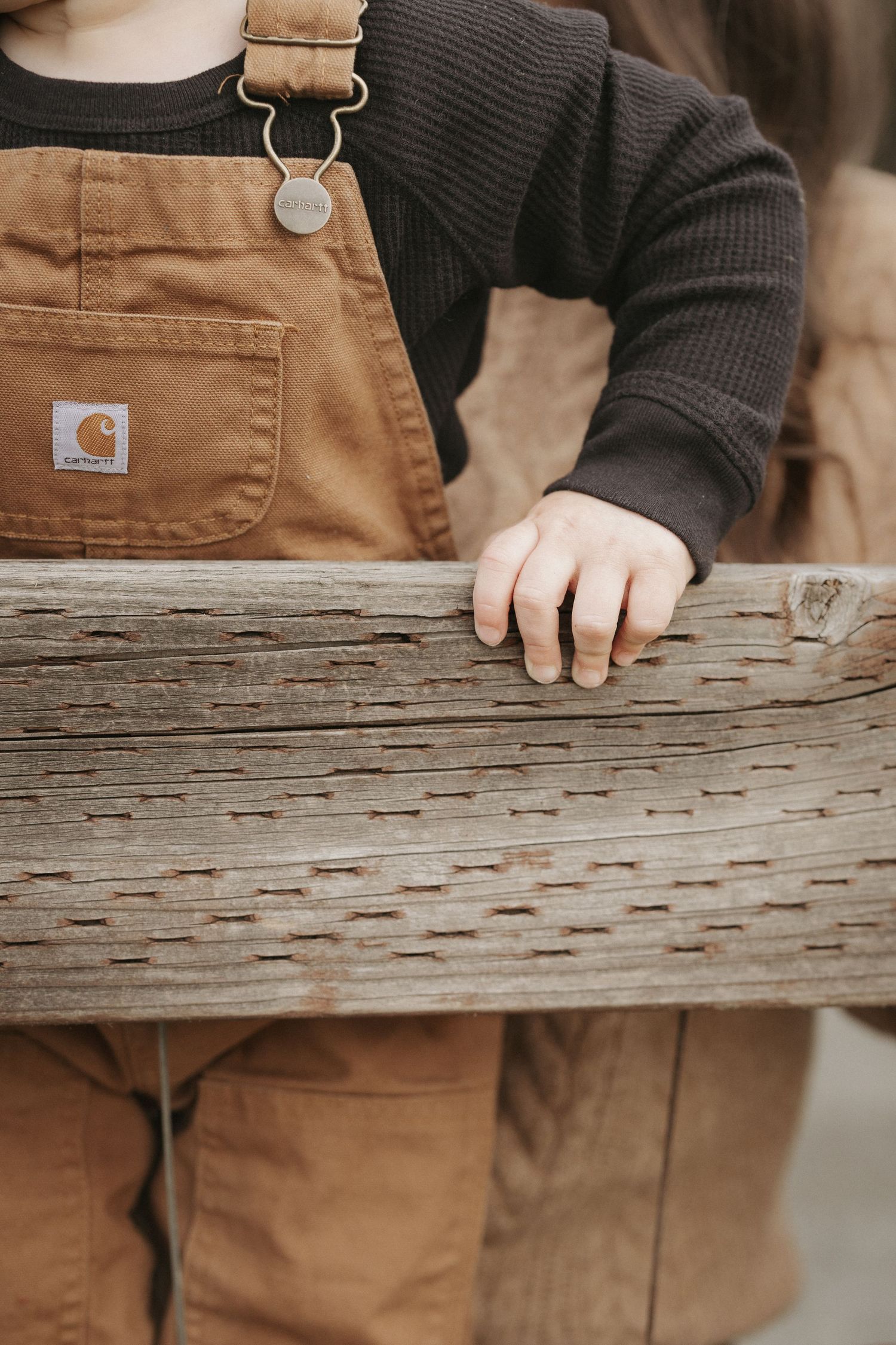 Child in Carhartt brown overalls and black shirt resting hand on weathered wooden fence rail.