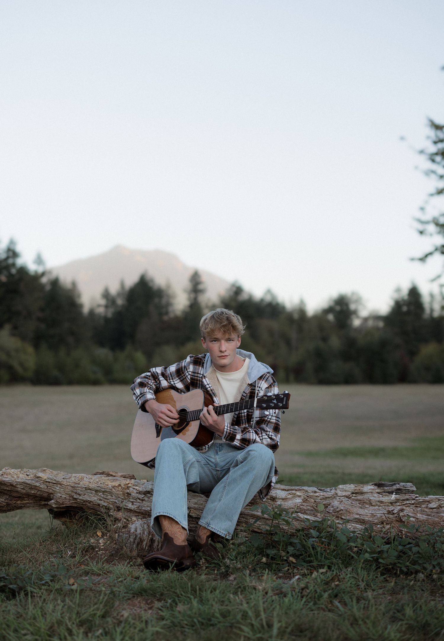 Young man playing acoustic guitar on a log in a scenic field with mountains and trees in the background.