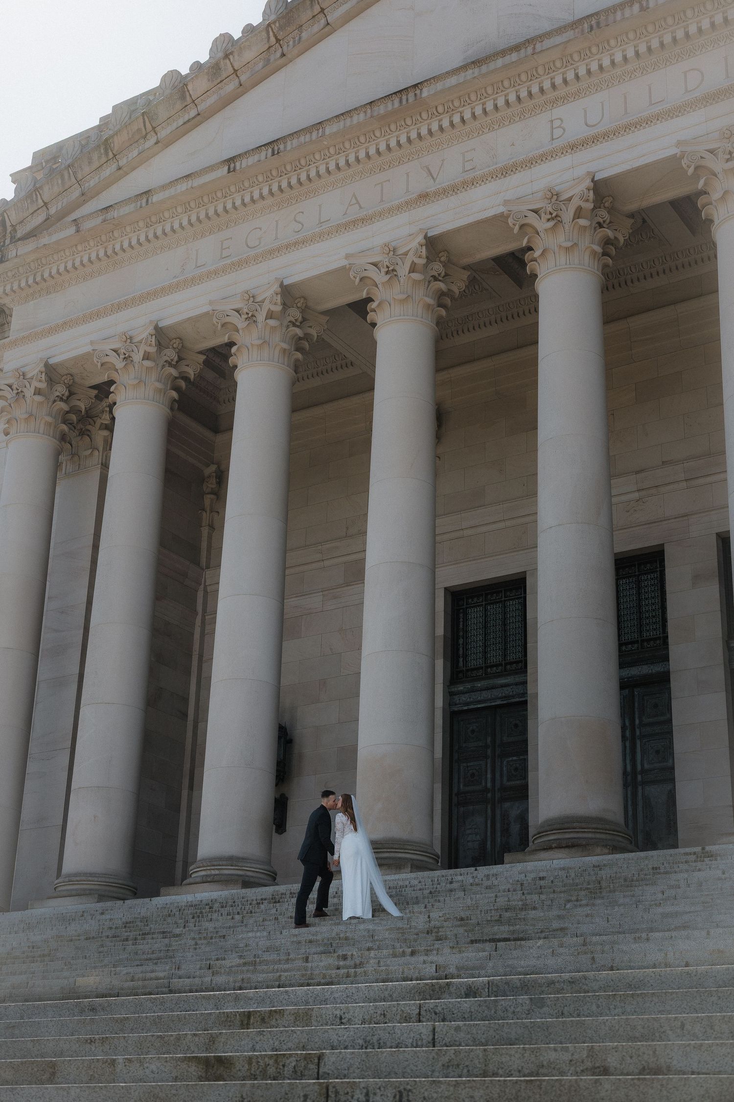 Bride and groom embrace on steps of grand neoclassical building with tall columns at their wedding.