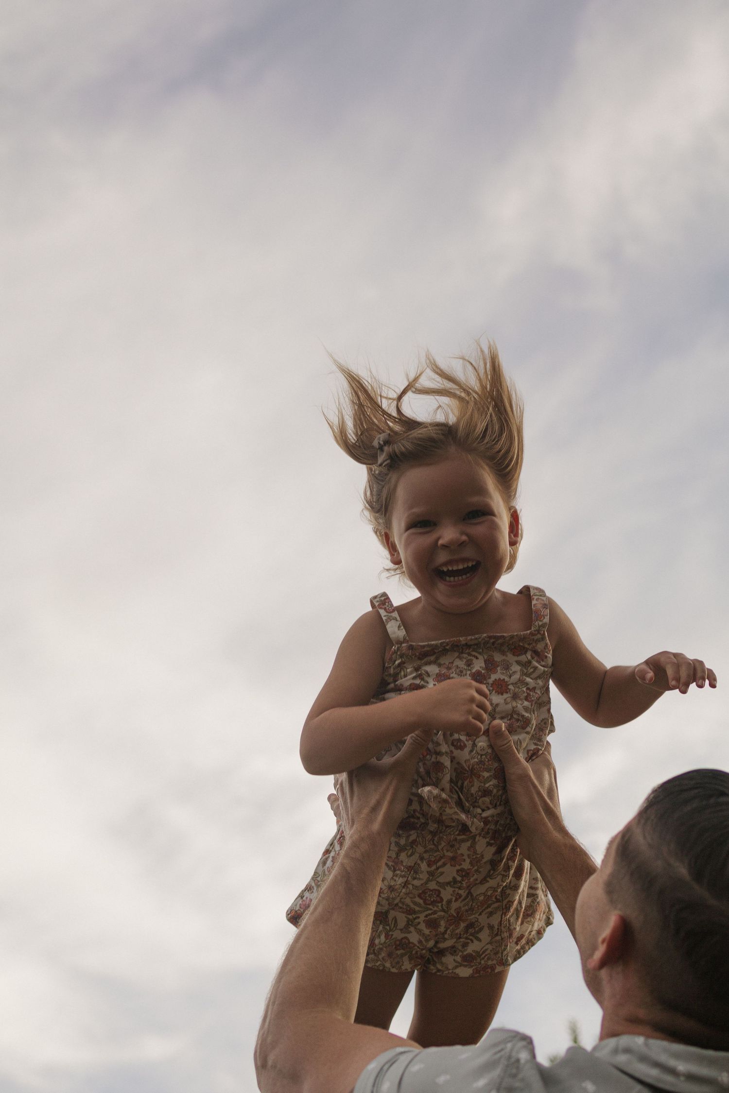 A joyful toddler girl is tossed in the air by a parent against a cloudy sky, her hair flying and laughing.