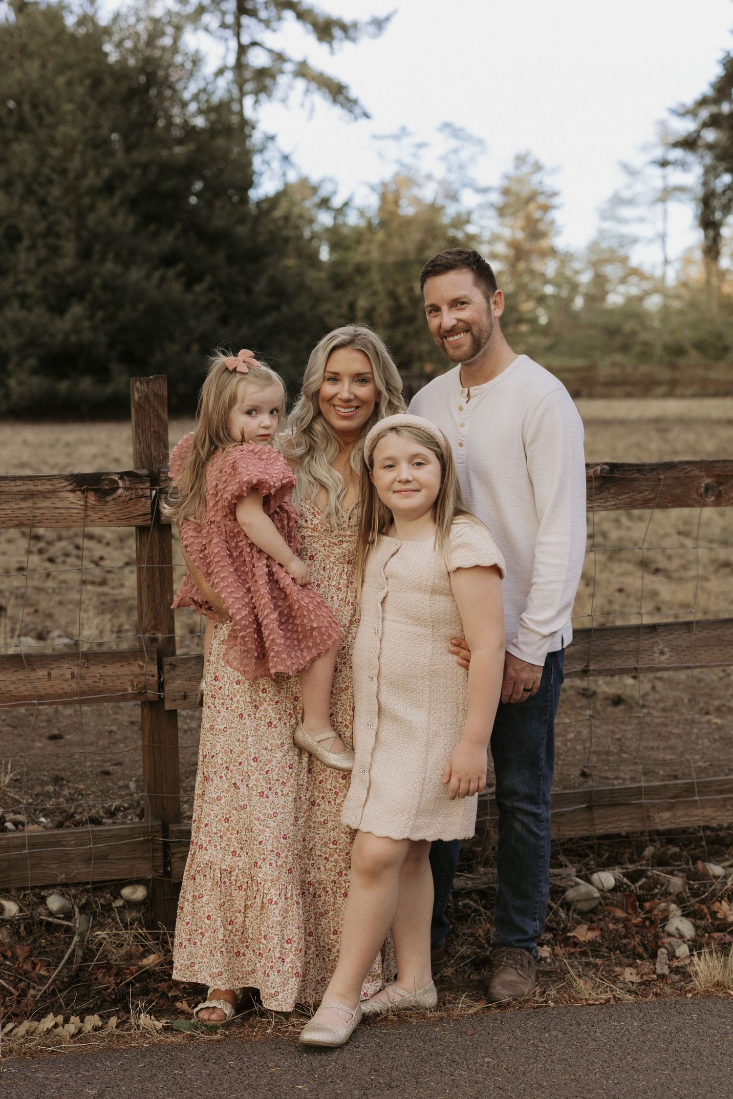 A smiling family of four poses outdoors by a wooden fence, surrounded by trees in a warm, natural setting.