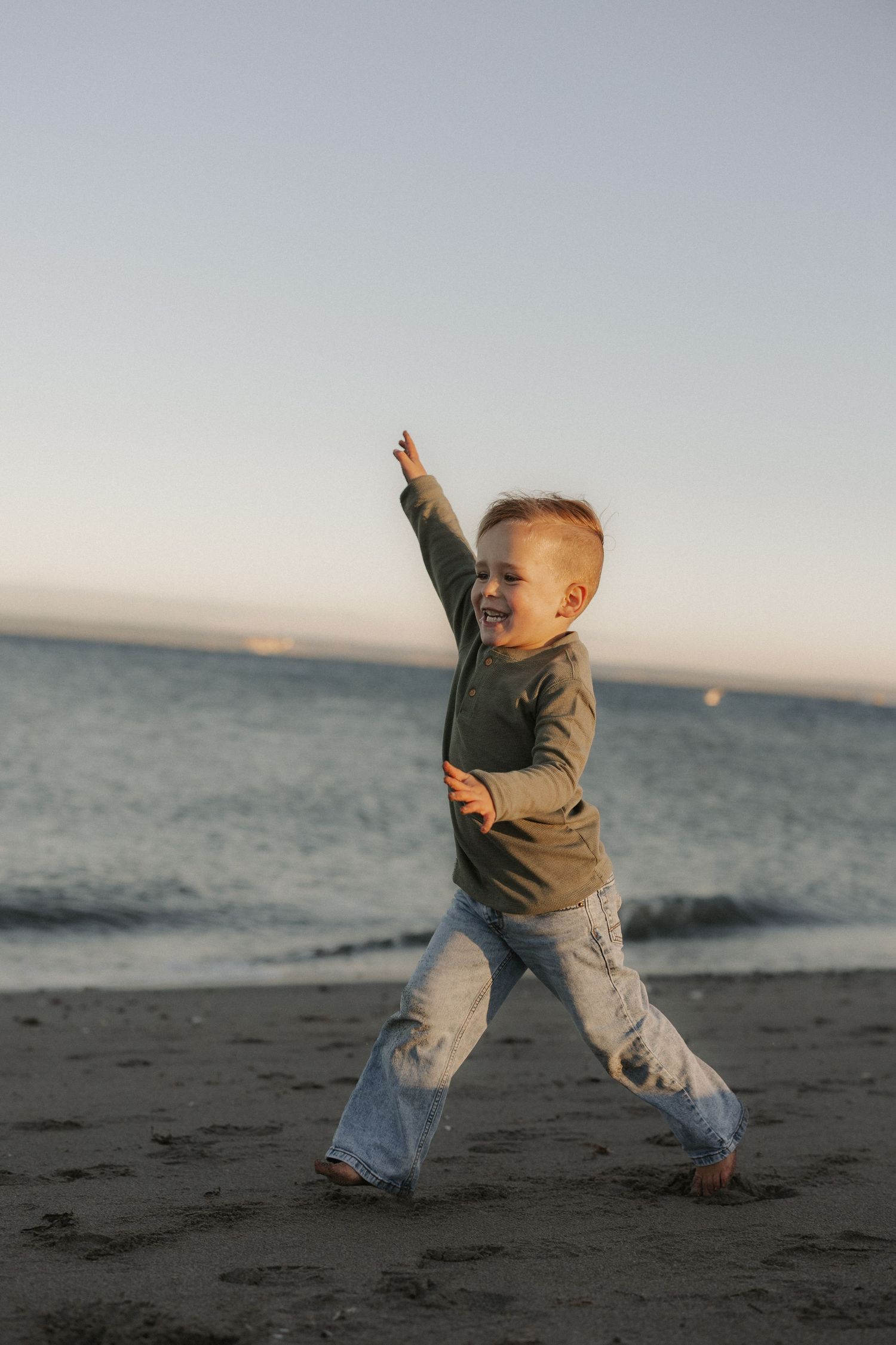 A joyful toddler plays on a sandy beach at sunset, raising one arm up while smiling.