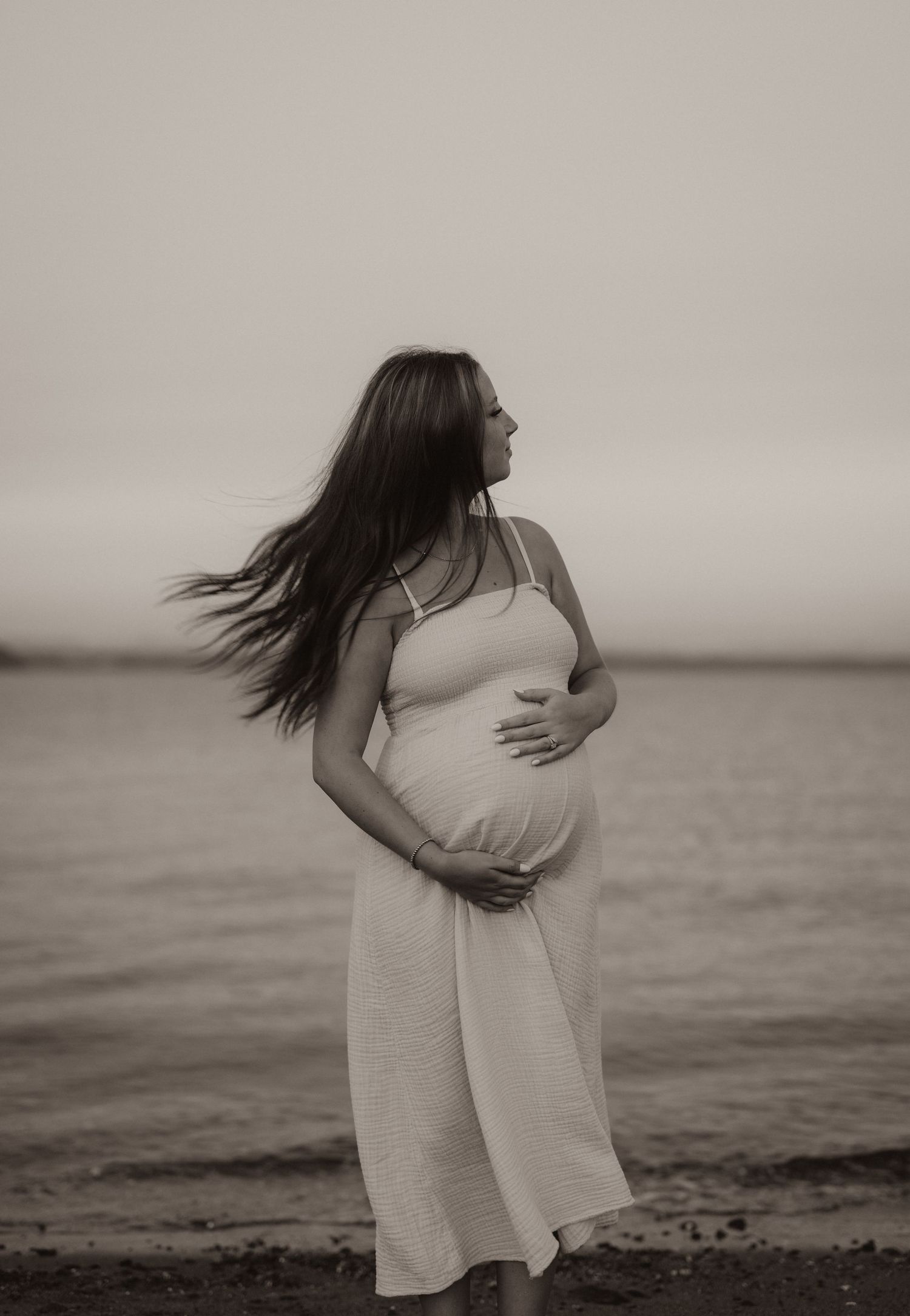 Pregnant woman in white dress holding belly at beach, black and white maternity photo with wind-blown hair.