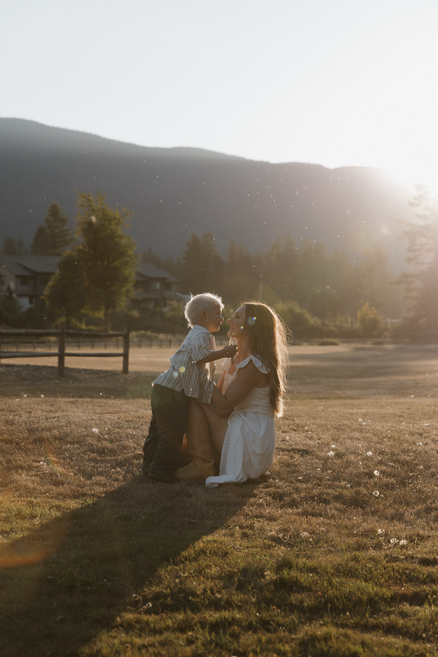 Mother and child share a tender kiss in golden sunset light with mountain backdrop in a scenic field.