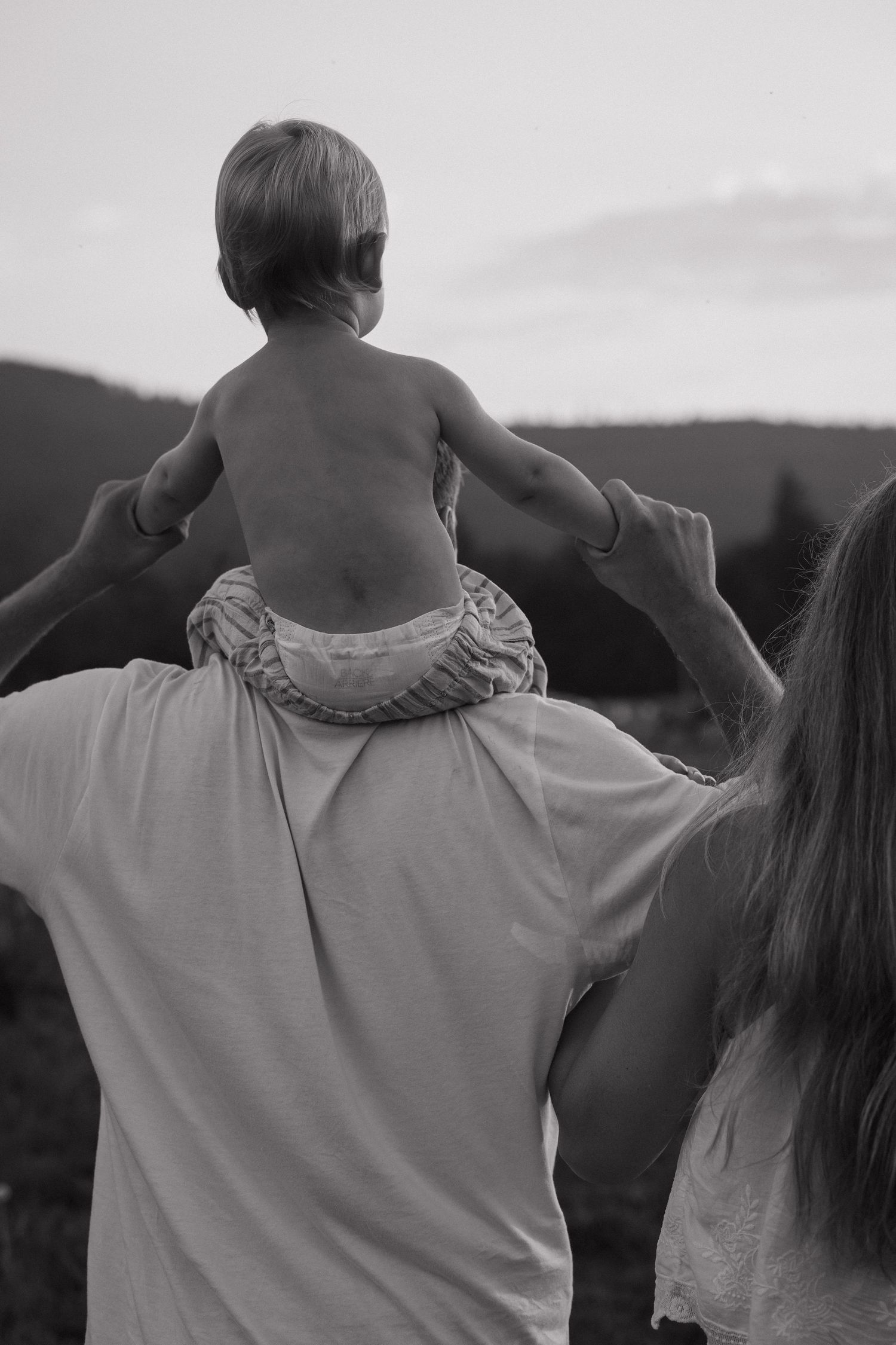 Black and white photo of parents holding toddler on shoulders outdoors with mountain landscape at dusk.