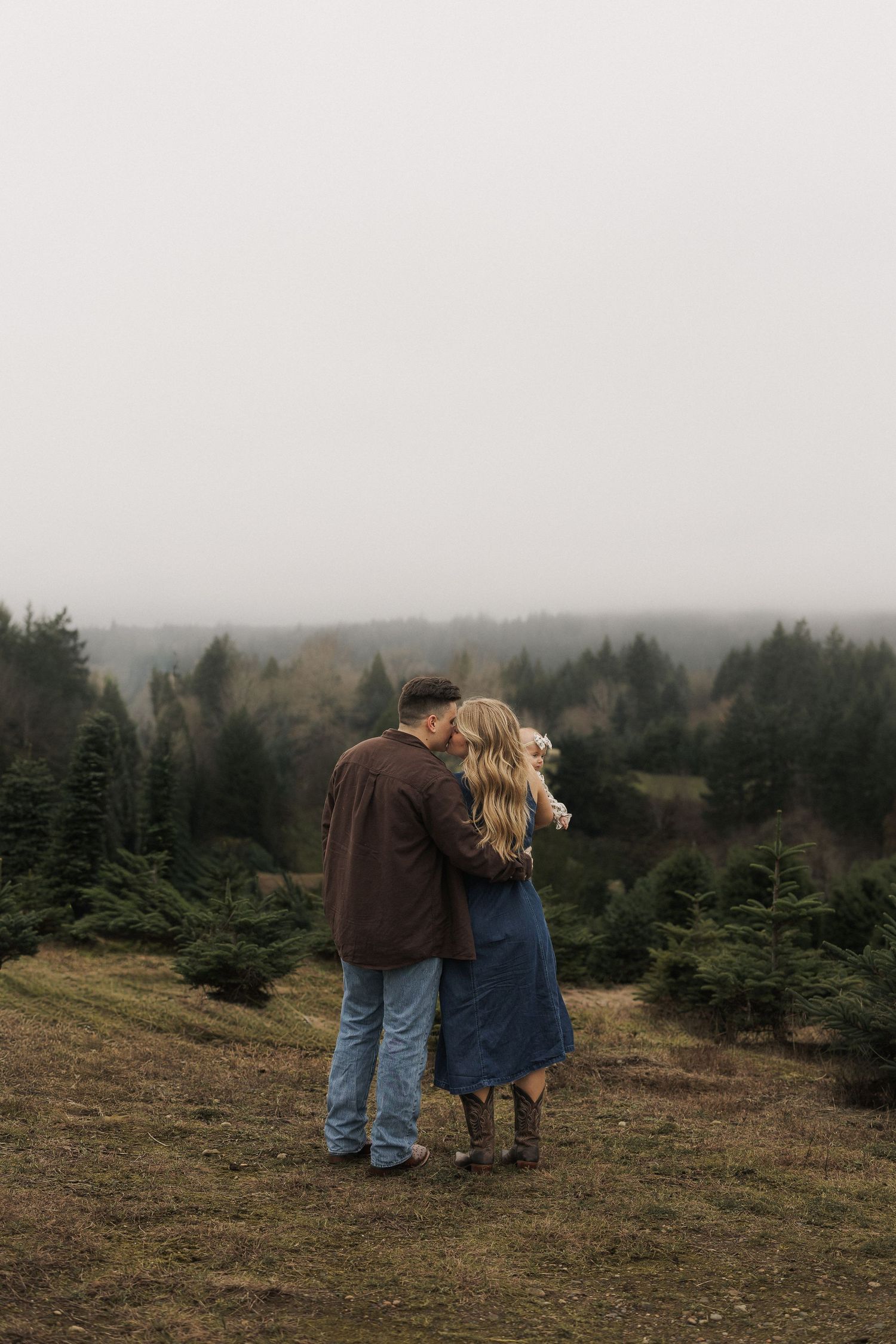Couple embracing on a misty hill overlooking a lush evergreen tree farm on a foggy day.