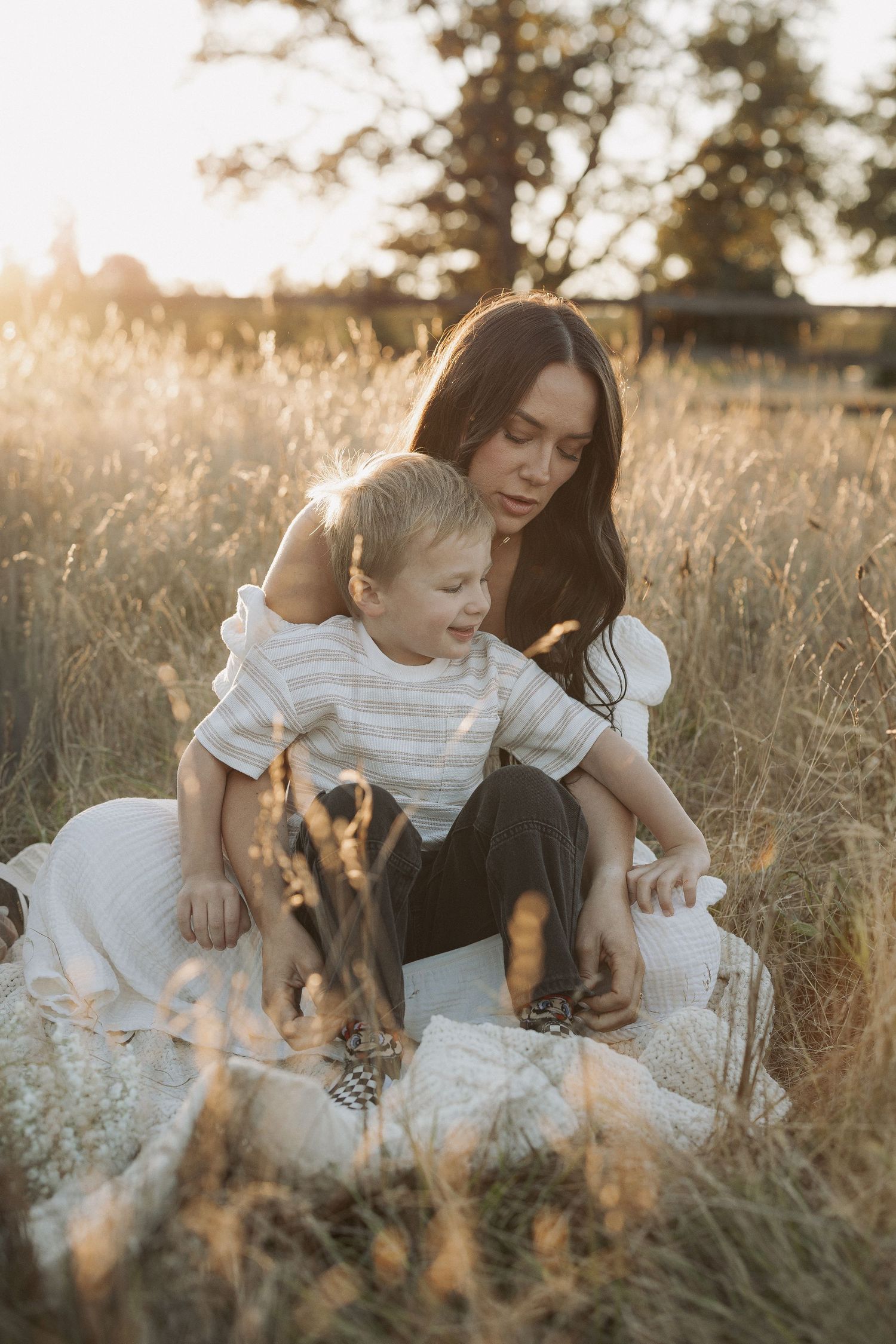 A mother and young son sit together on a blanket in a golden sunlit field during sunset.