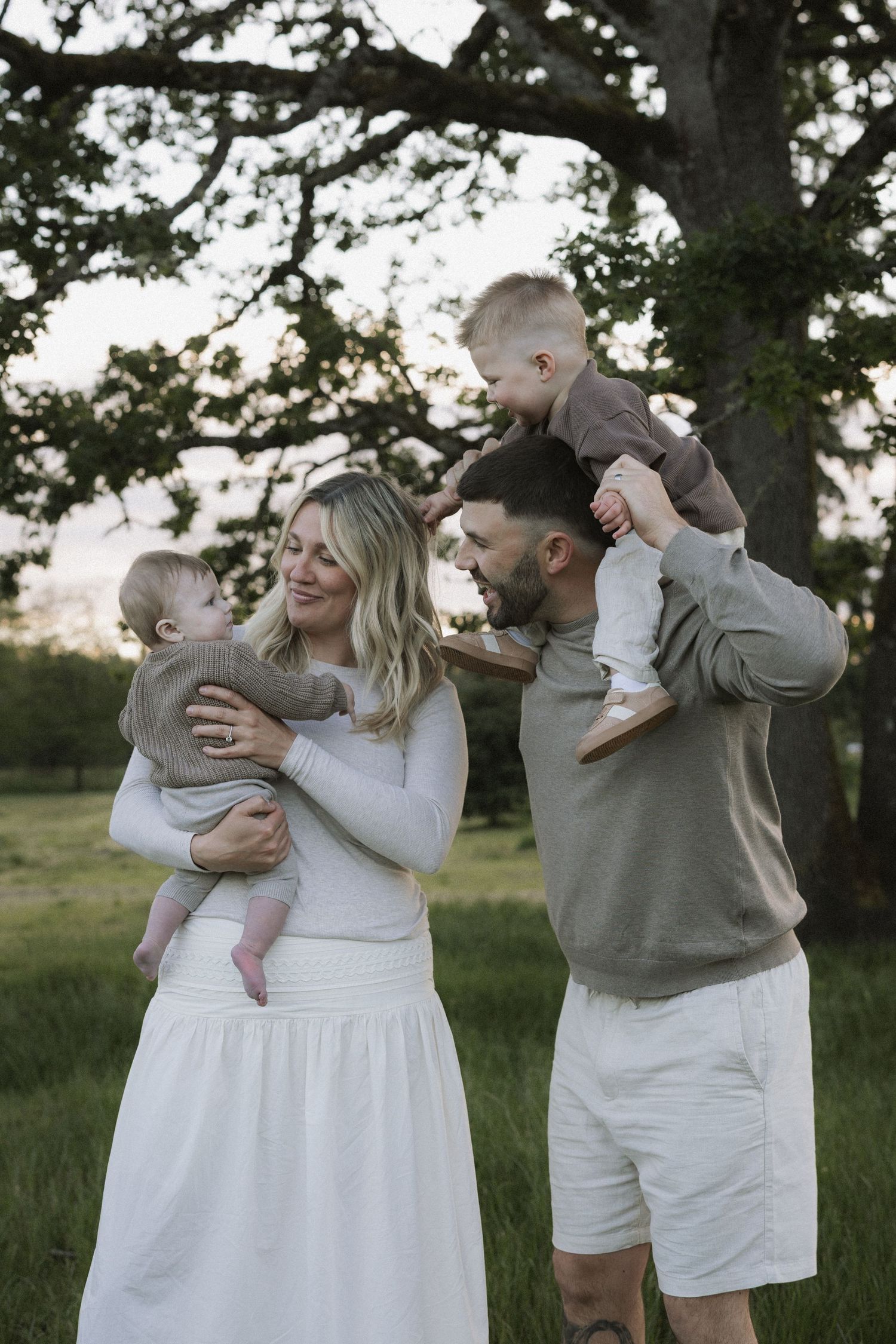 A happy family of four poses outdoors, with a woman in white holding a baby and a man carrying a toddler on his shoulders.