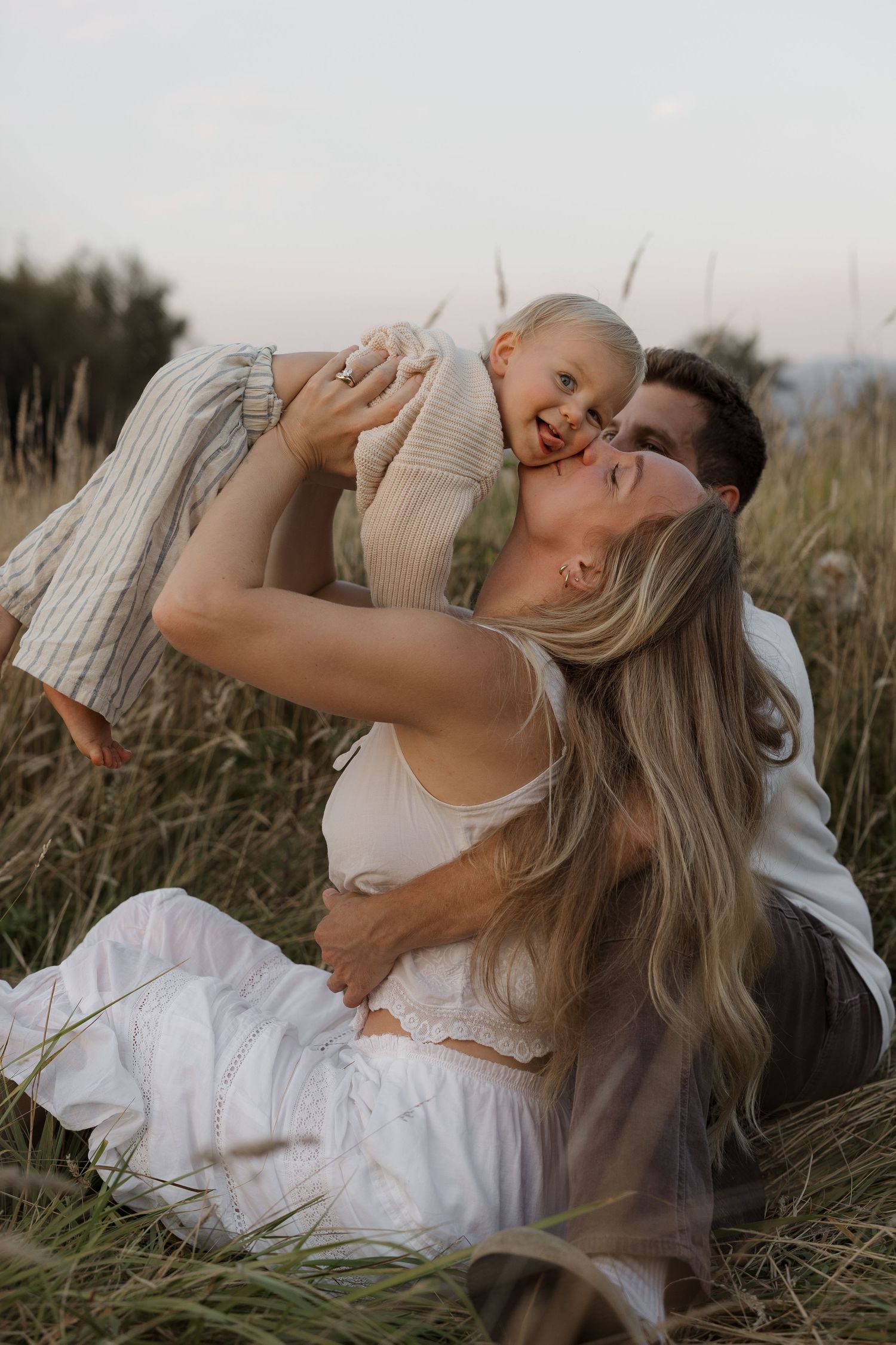 A happy family sits in a golden field at dusk, mom lifting a laughing toddler while dad embraces them both.