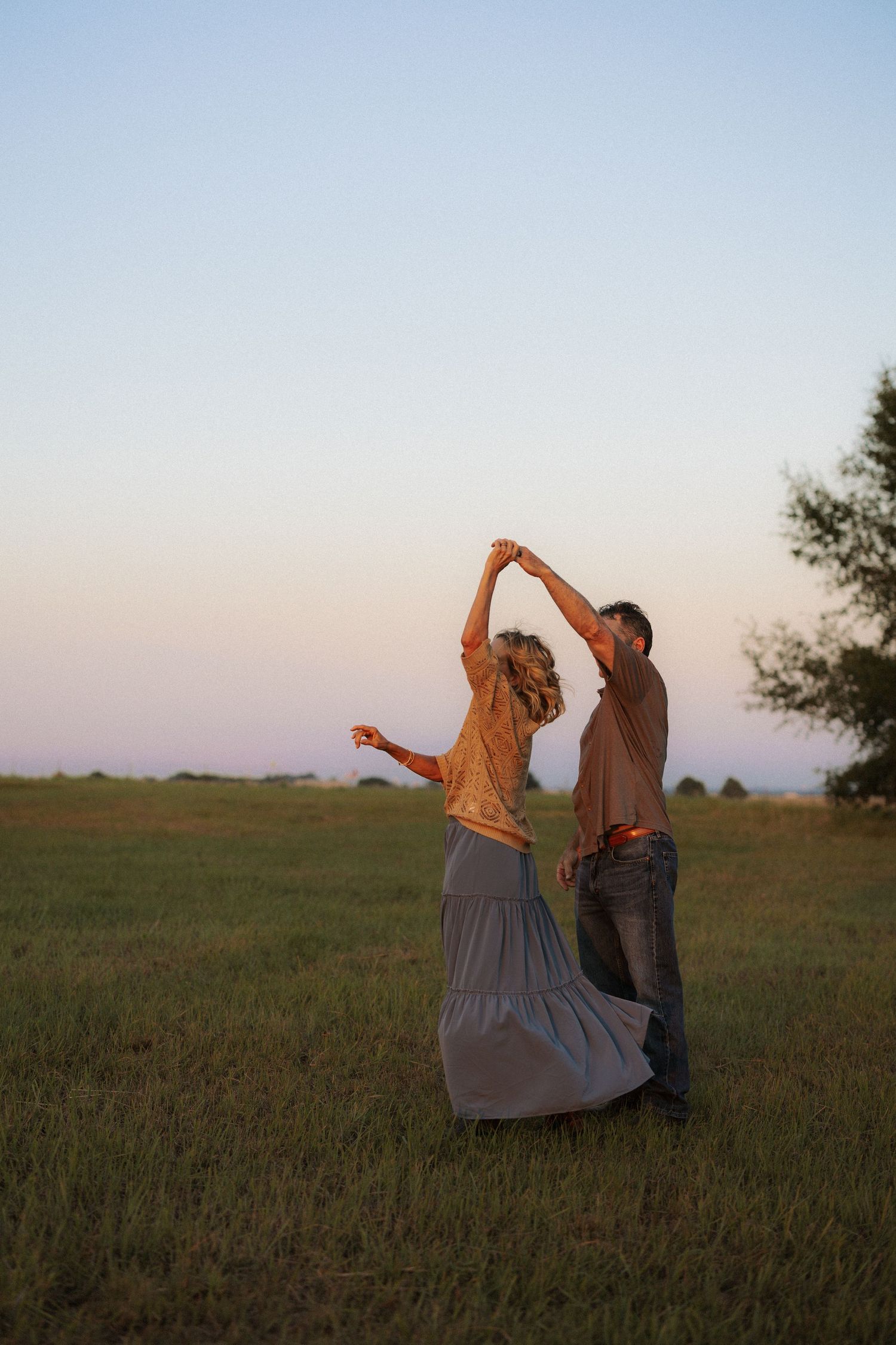 Couple dancing and spinning joyfully in an open grassy field at golden hour sunset.