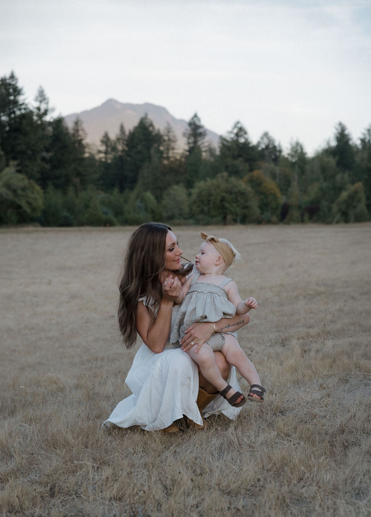 A mother in a white dress crouches in a golden field, lovingly holding her baby girl with mountains and trees behind.