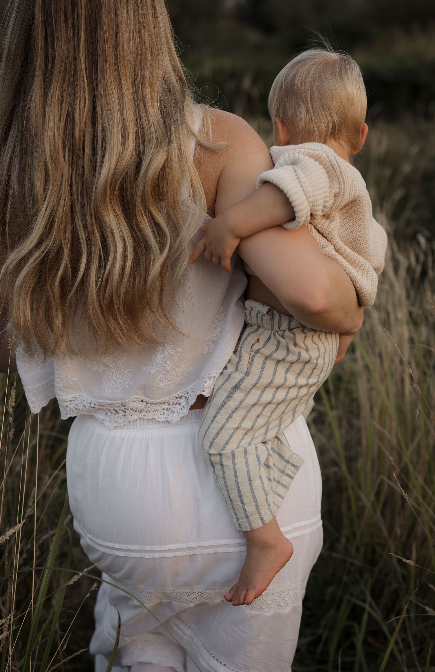 Mother with long blonde hair holding barefoot baby in neutral knit outfit in tall grass.