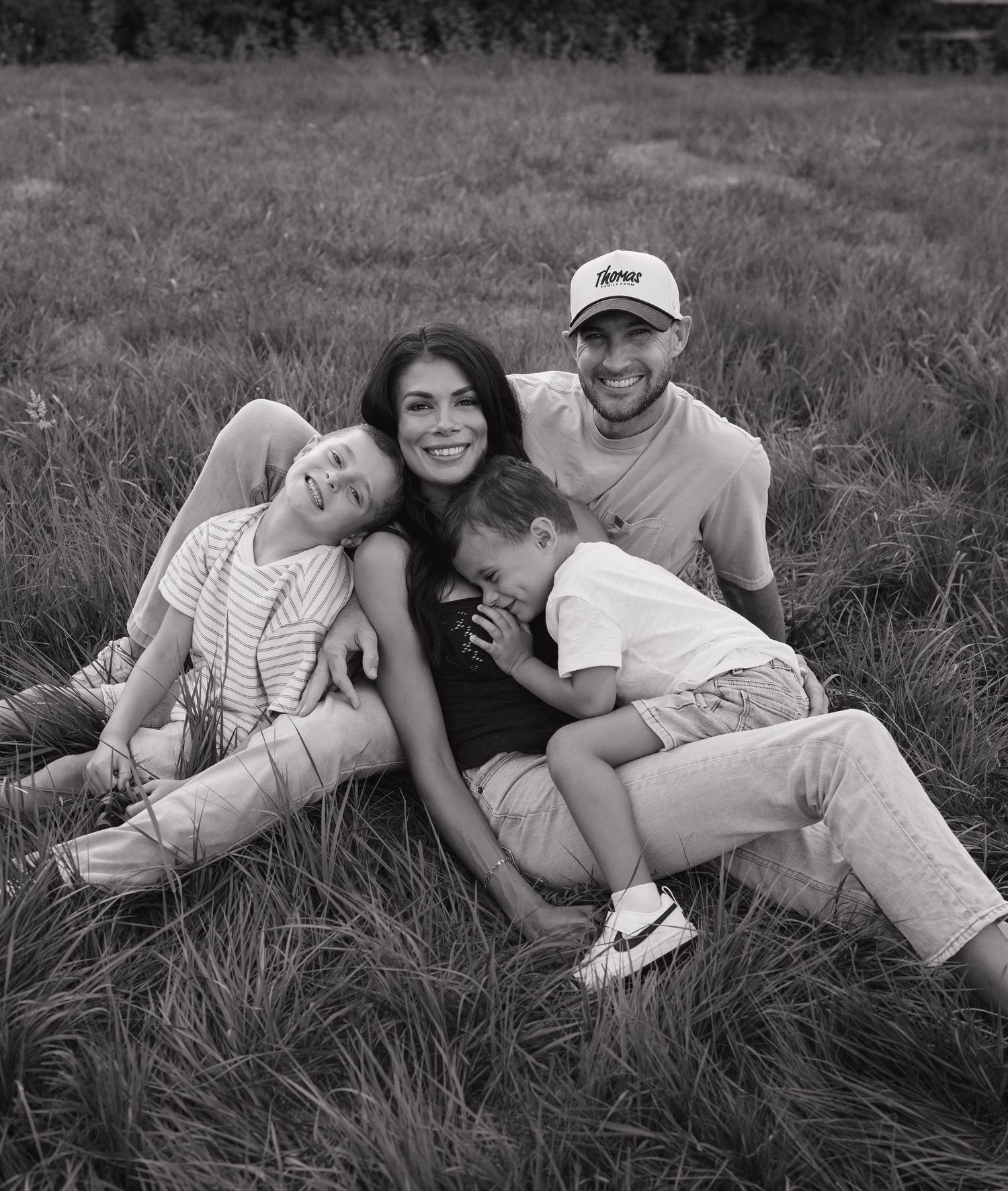 Black and white photo of a happy family of four sitting together in a grassy field, smiling and embracing outdoors.