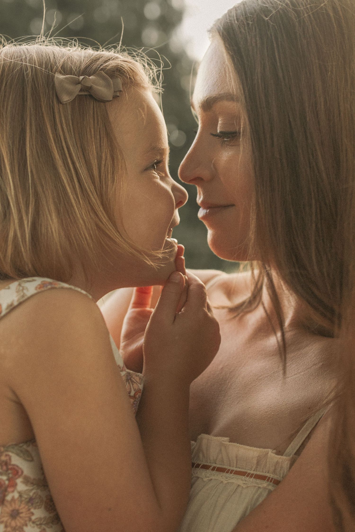A mother and young daughter share a tender nose-to-nose moment in warm golden sunlight.