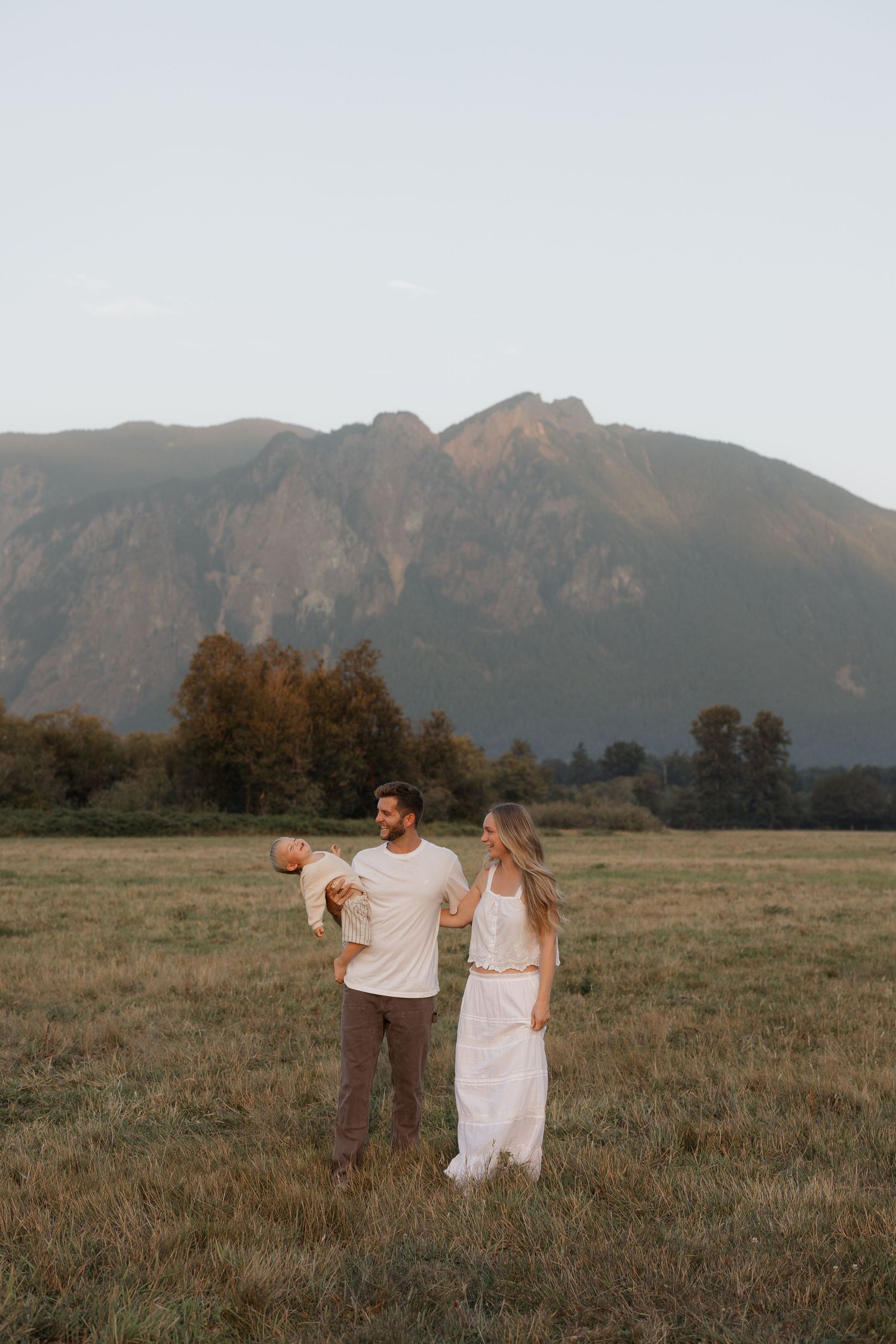 A couple stands in a mountain meadow at golden hour, man holding a baby while woman in white dress looks on adoringly.