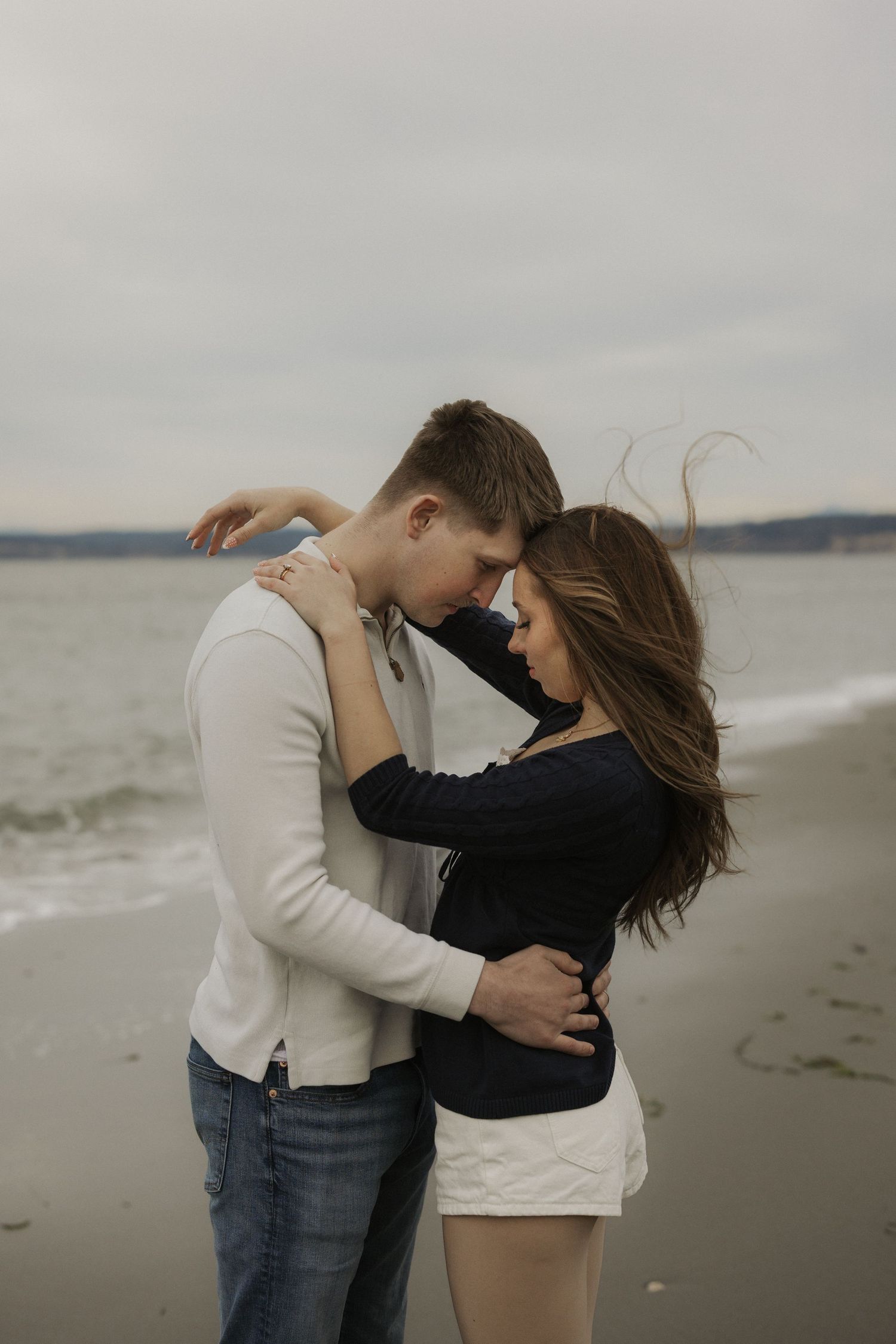 Romantic couple embracing on a cloudy beach, woman in black top and white shorts, man in white sweater and jeans.