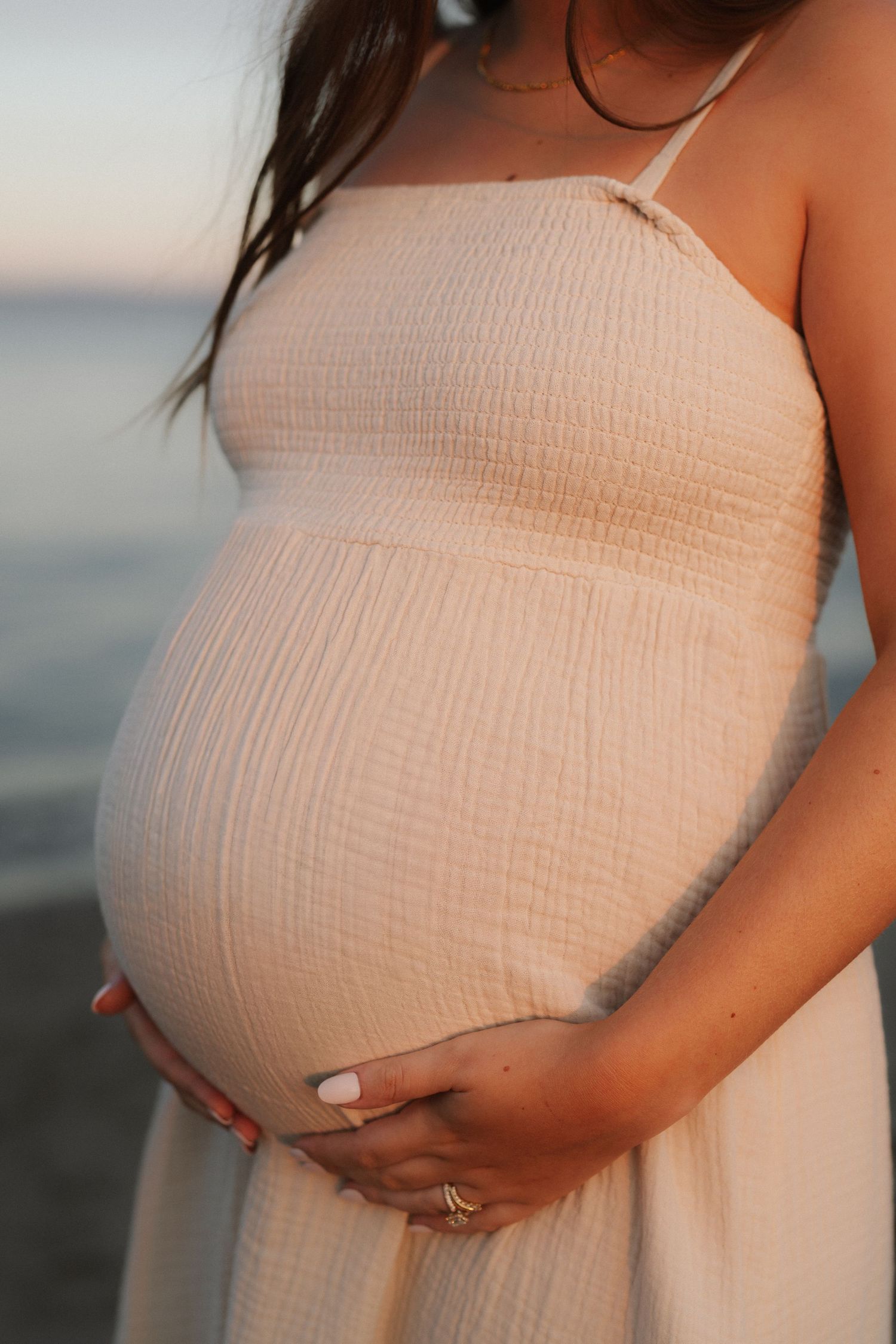 Close-up of a pregnant woman in a cream dress gently holding her bare belly near the ocean at sunset.