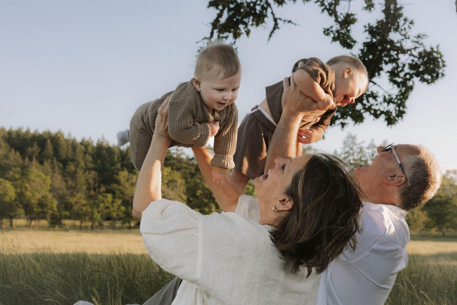 Happy parents raising twin babies up in a sunlit outdoor meadow setting.